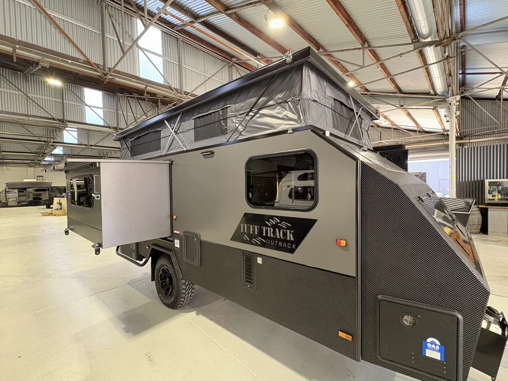 A gray off-road camper trailer named 'Tuff Track Outback' with a pop-up roof, inside a warehouse.
