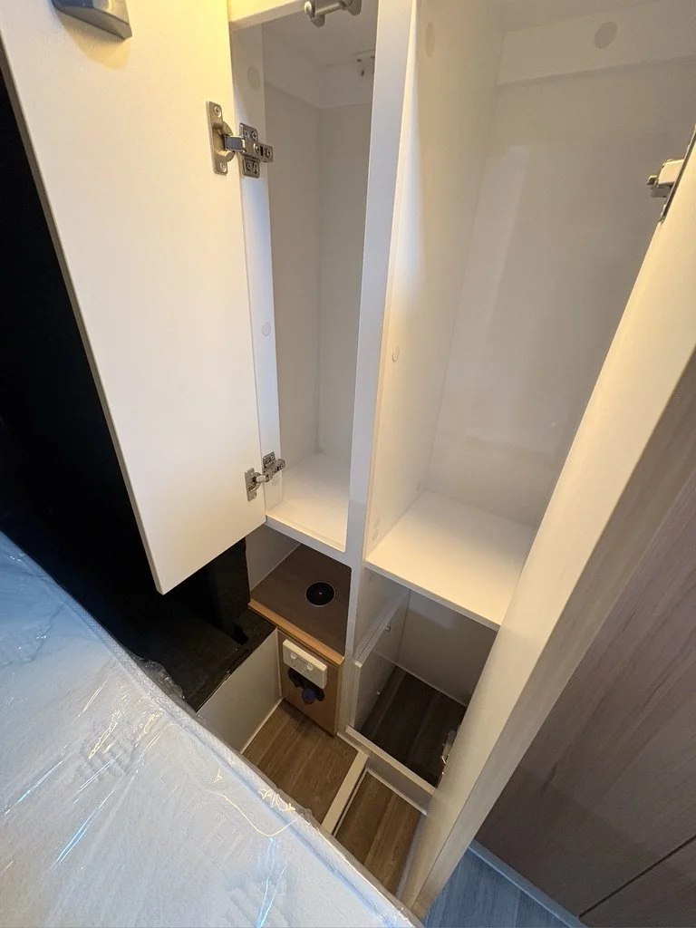 Open cabinet with empty white shelves, a small cabinet with a power outlet and a circular cutout, situated next to a bed with plastic protective cover.