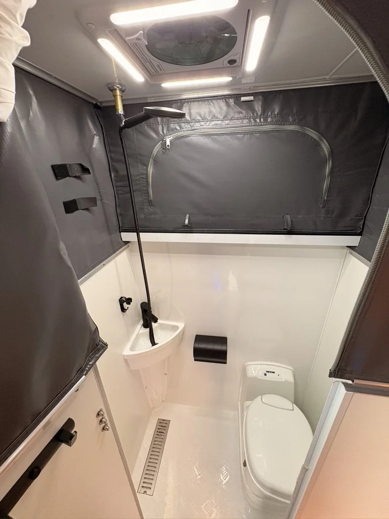 Compact bathroom with a showerhead, a toilet, a small sink, and black privacy curtains designed for a mobile or RV space.