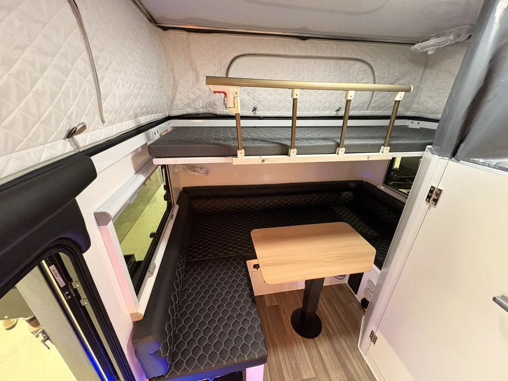 Interior of a small living space with a bunk bed and a small table. The bunk bed has a sleeping area on top with a safety rail and a mattress, and a seating area below with a black padded cushion. There's a window to the left and a small door to the 