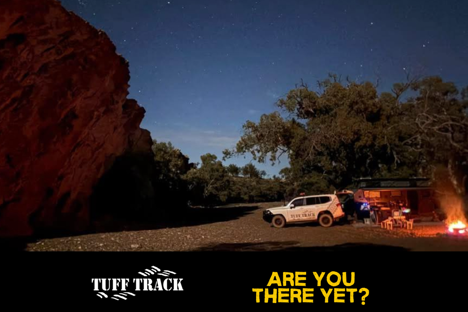A nighttime outdoor scene with a campfire, a white off-road vehicle labeled 'TUFF TRACK,' and a group of people beside a small cabin or shelter surrounded by trees under a starry sky.