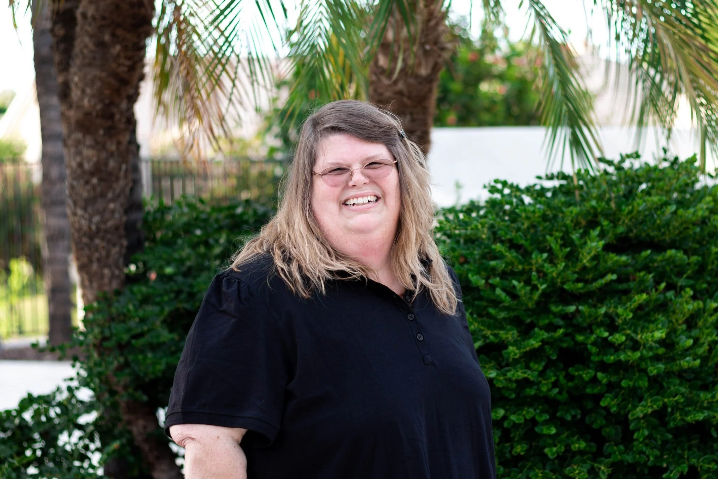 A woman with shoulder-length blonde hair, glasses, and a black shirt standing outdoors in front of green bushes and palm trees and smiling.