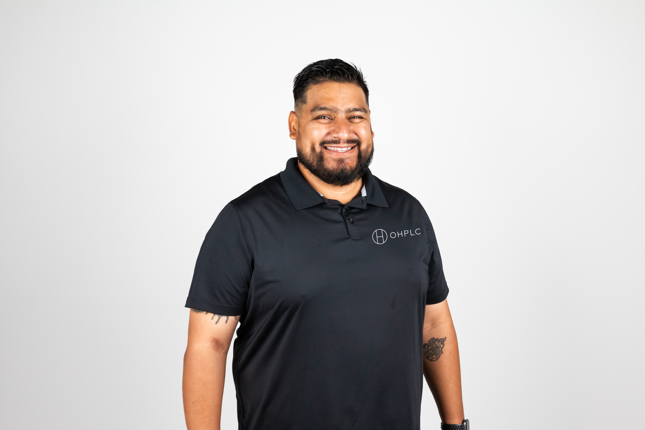 A smiling man with a beard and tattoos wearing a black OHPLC polo shirt against a plain white background.
