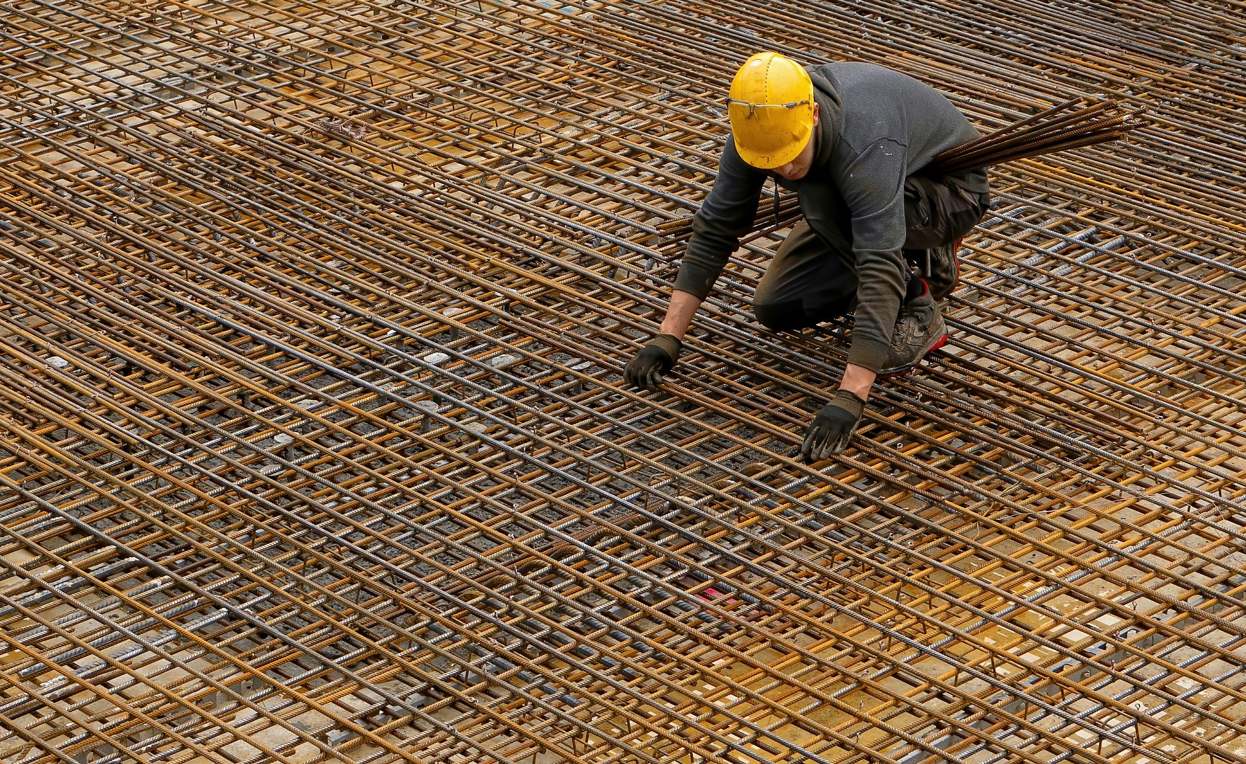 Construction worker in a yellow safety helmet installing or inspecting steel rebar on a concrete building foundation.