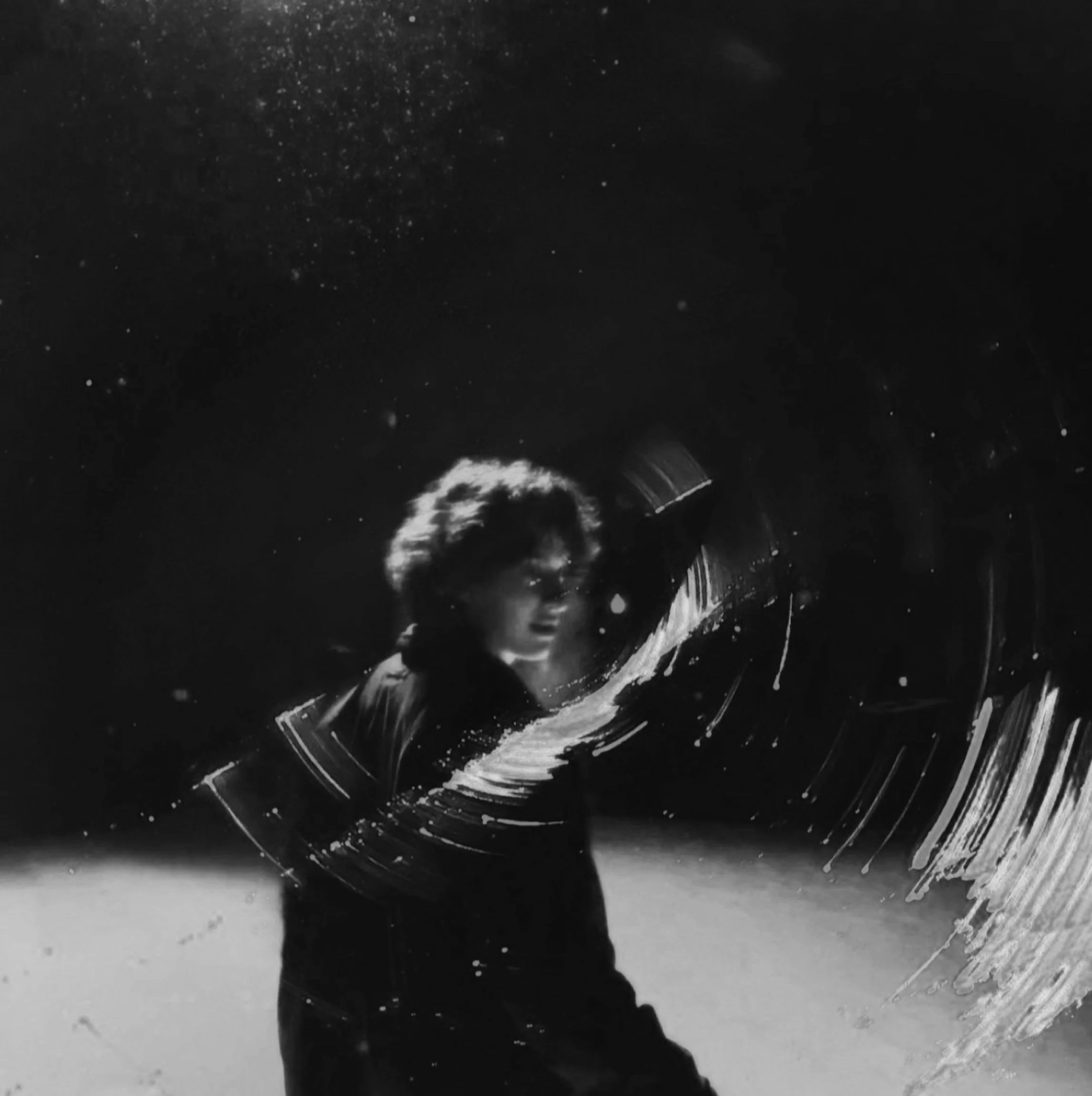 A person with curly hair and glasses, drawing with a large brush on a glass surface, creating sweeping strokes in a dark setting.