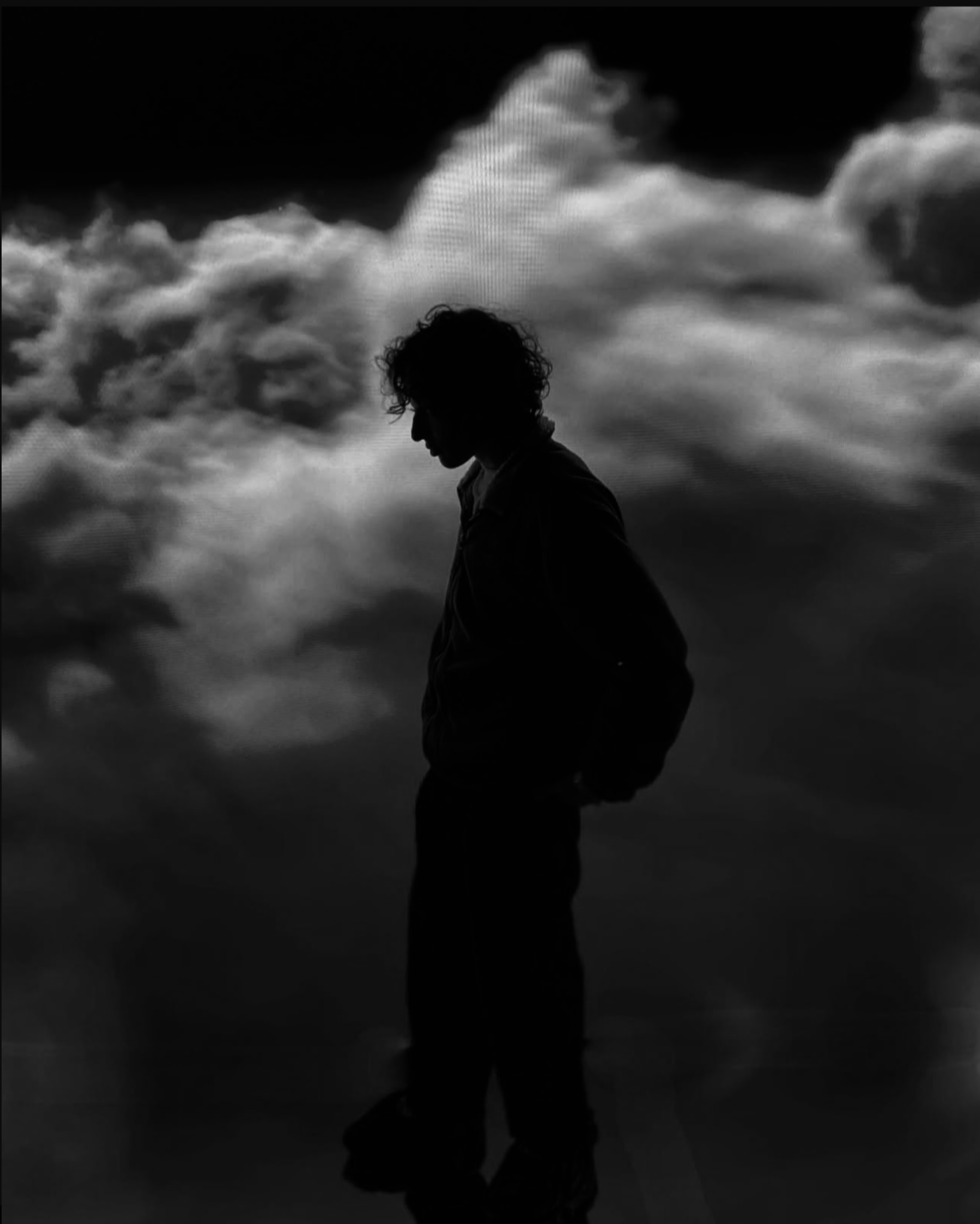 Silhouette of a person with curly hair standing with head bowed against a backdrop of cloudy sky
