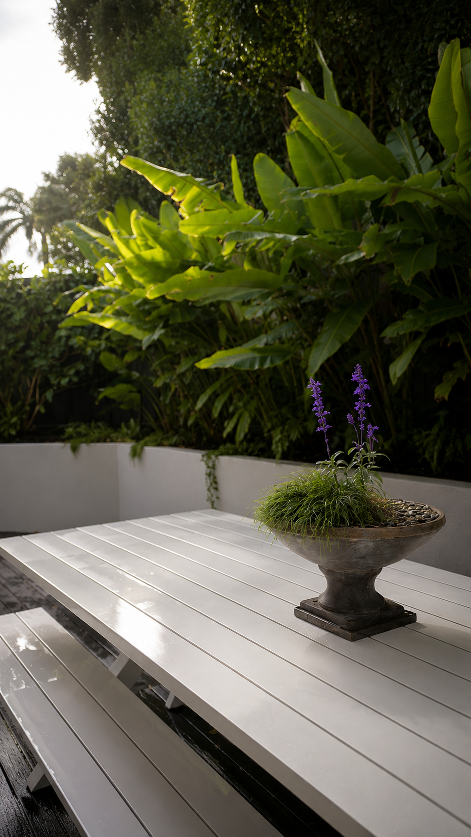 Outdoor dining table positioned beside lush tropical screening plants and raised rendered planters, creating a private backyard entertaining space with layered planting.