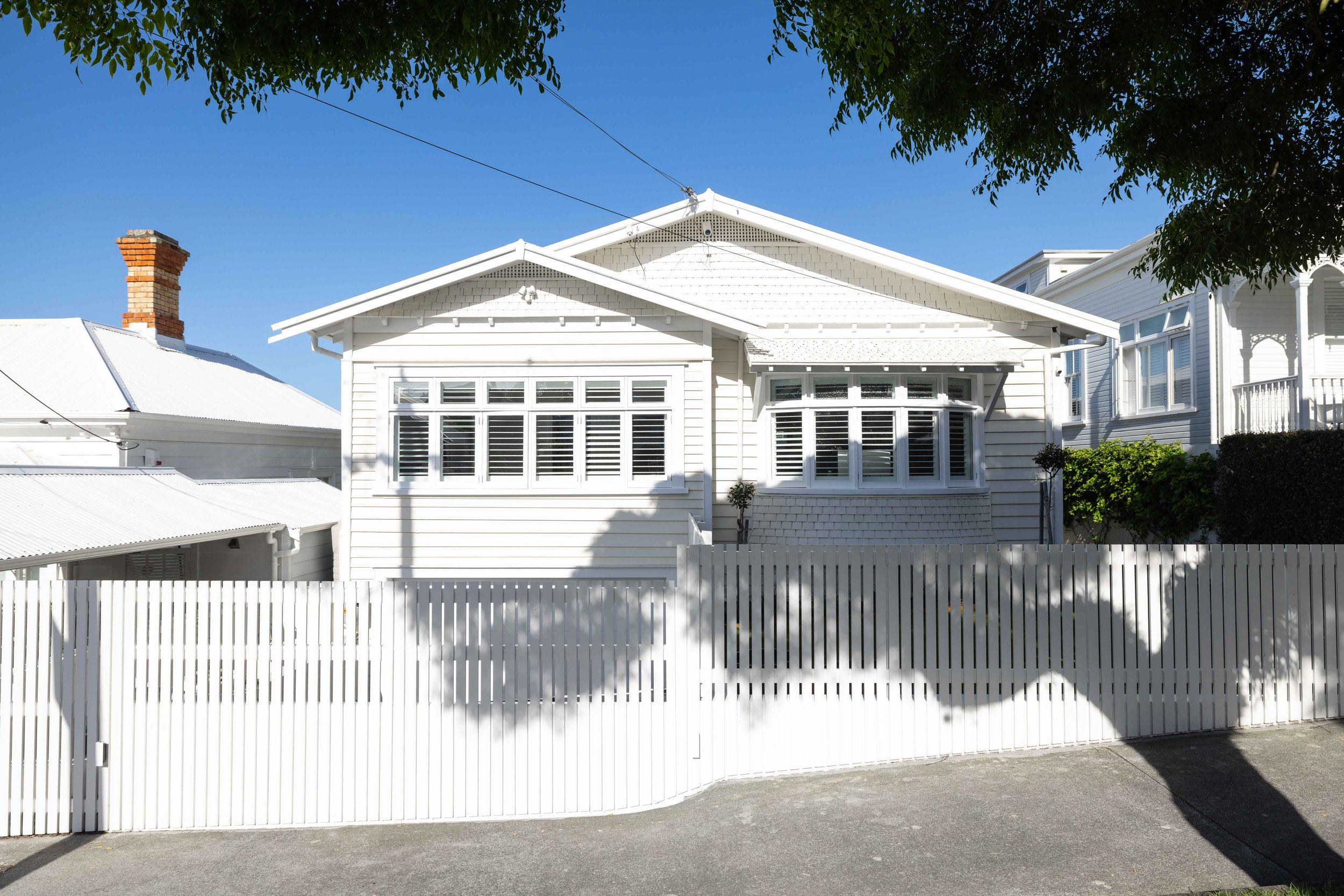 White house with large bay window, white picket fence in front, and trees overhead against a bright blue sky.