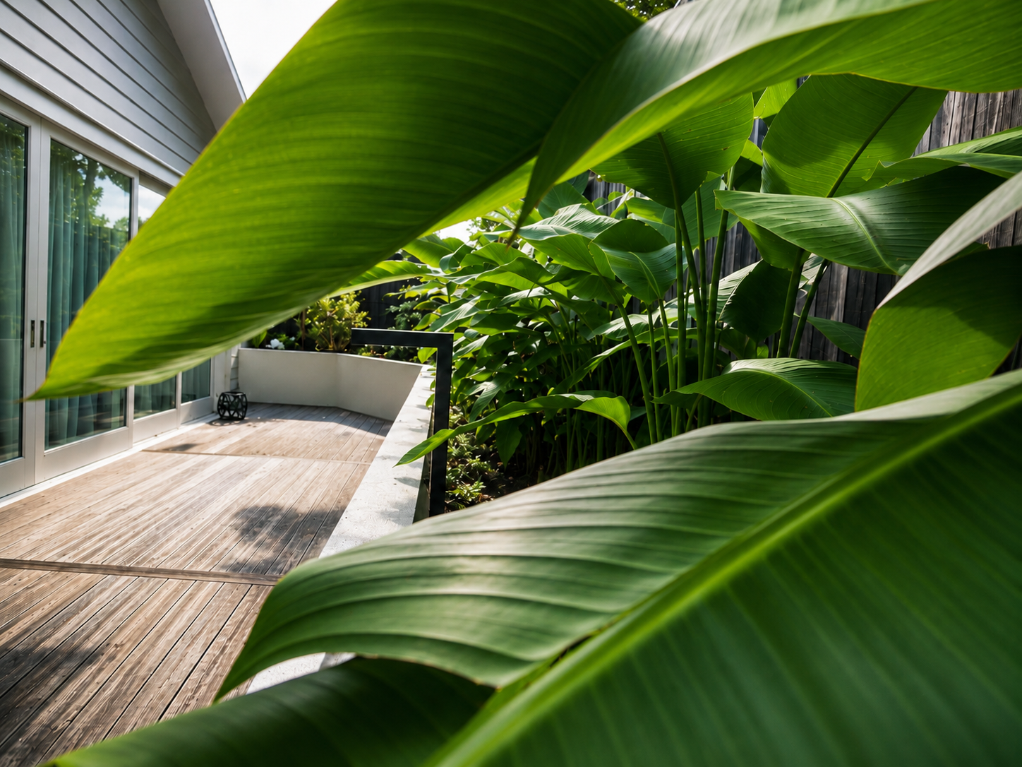 Modern outdoor entertaining area with dense tropical screening, raised planter beds, timber decking, and a white picnic-style table in a landscaped residential garden.
