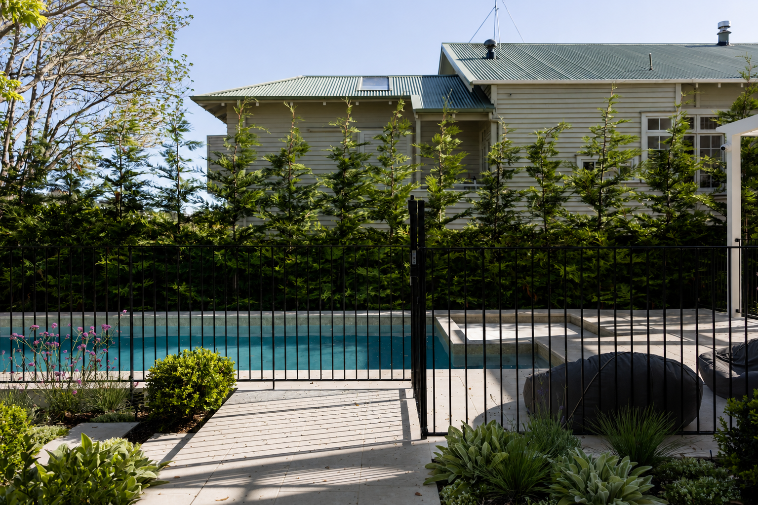 Modern black steel pool fencing surrounding a landscaped swimming pool with natural stone paving, feature planting, and stone wall in Auckland, New Zealand.