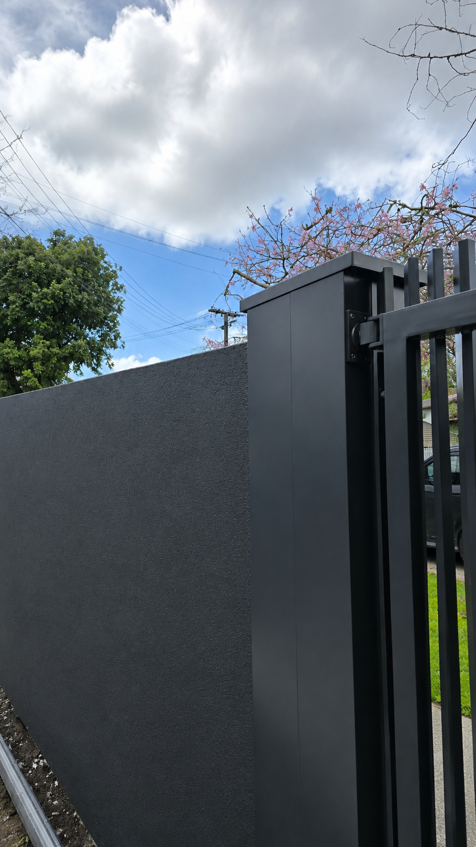 Close-up of a charcoal rendered boundary wall with a matte black aluminium gate and architectural gate post at a modern Auckland residential property.