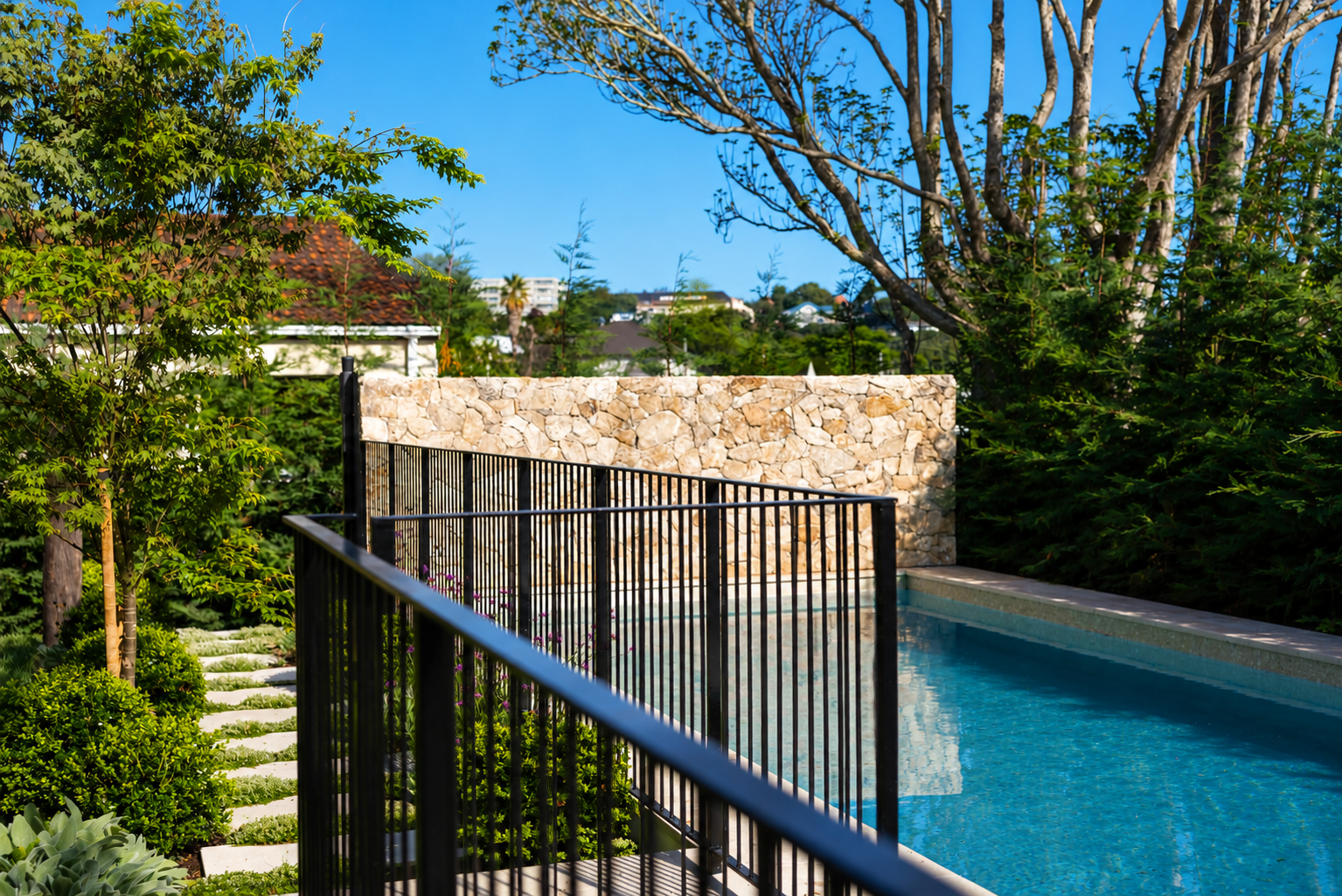 Modern black steel pool fencing surrounding a luxury swimming pool with landscaped gardens, natural stone feature wall and stepping stone pathway in Auckland, New Zealand.