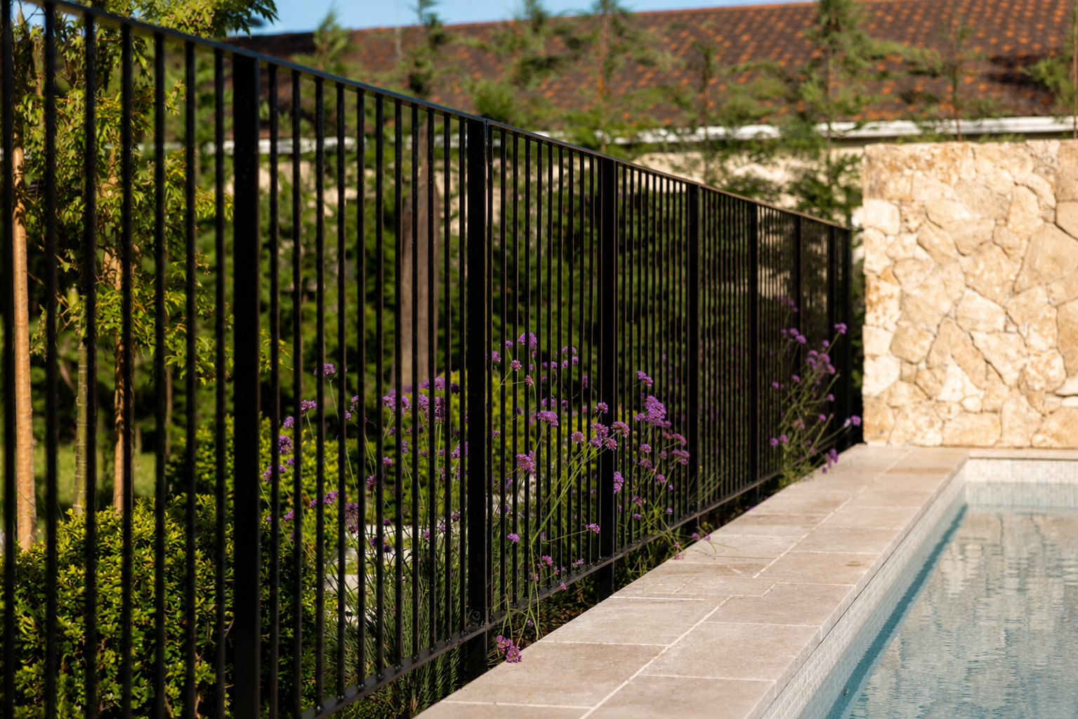 Close-up of a modern black steel pool fence beside a natural stone swimming pool with landscaped planting and feature stone wall in Auckland, New Zealand.
