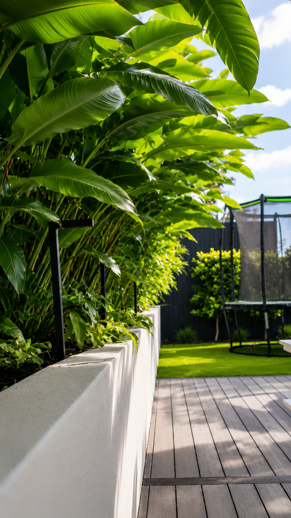 Modern outdoor entertaining area with dense tropical screening, raised planter beds, timber decking, and a white picnic-style table in a landscaped residential garden.