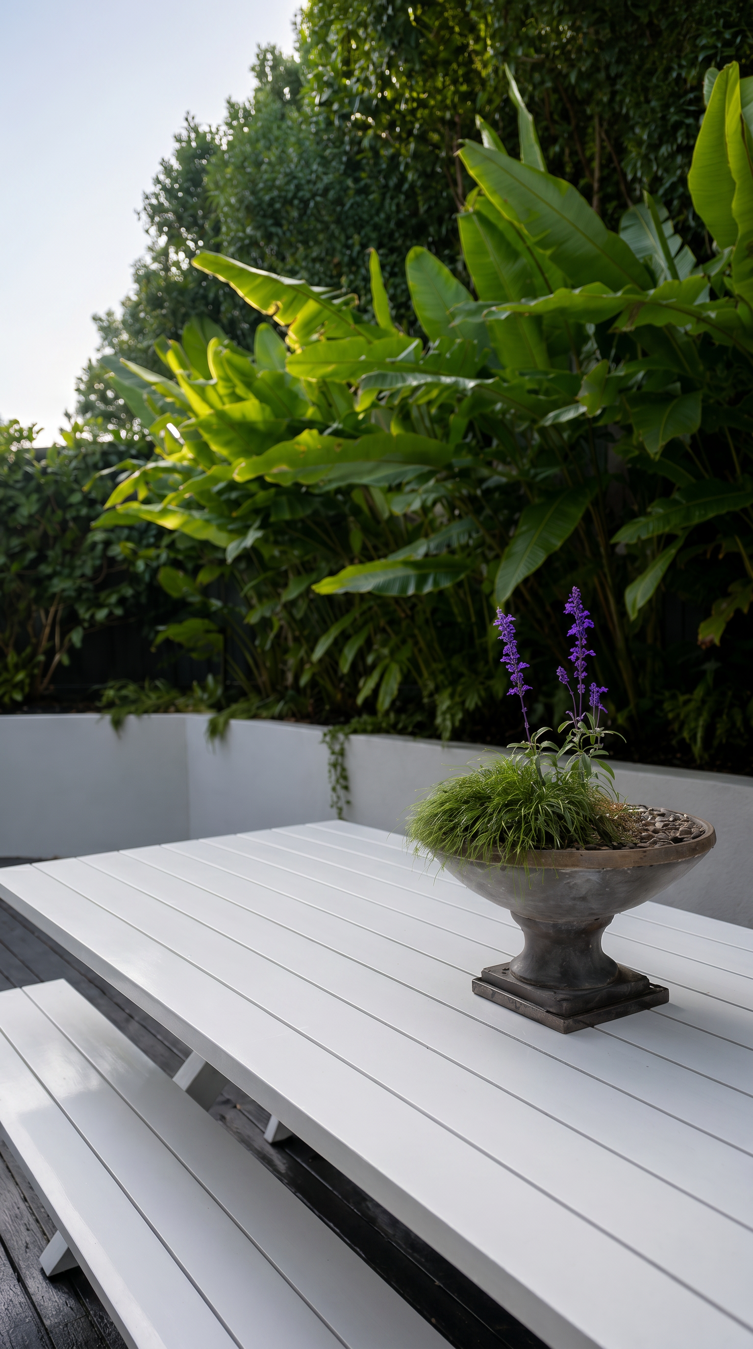 Outdoor dining table positioned beside lush tropical screening plants and raised rendered planters, creating a private backyard entertaining space with layered planting.