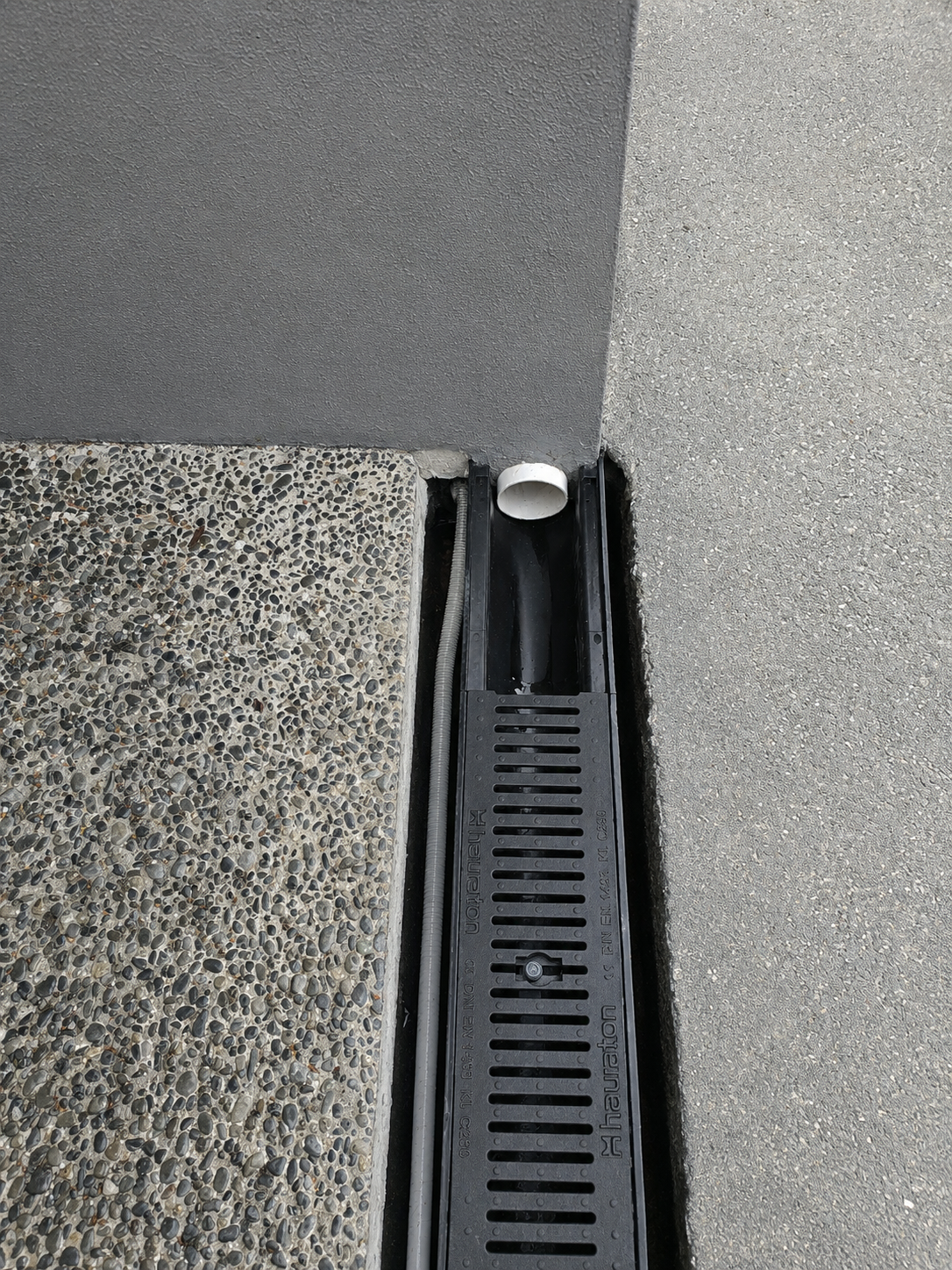 Linear channel drain installation at a residential property showing exposed aggregate concrete, asphalt and stormwater drainage detail completed by Whenua Scapes in Auckland.