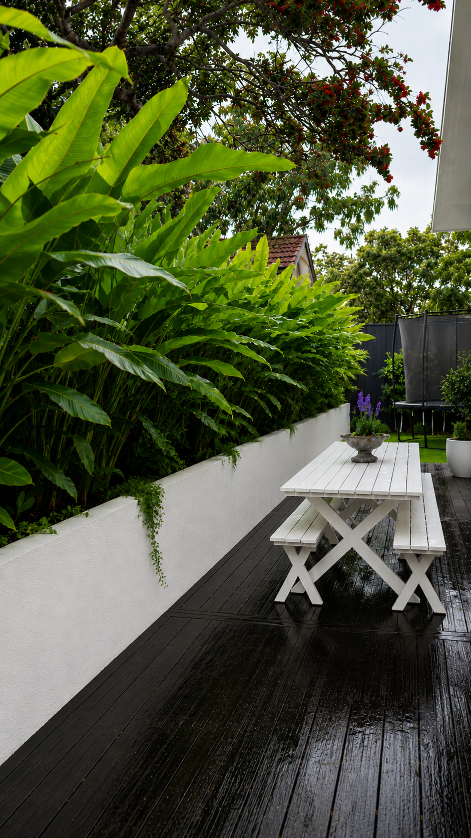 Contemporary backyard landscape featuring lush tropical planting, white rendered retaining walls, a white outdoor dining table, and wet timber decking creating a private outdoor entertaining space.