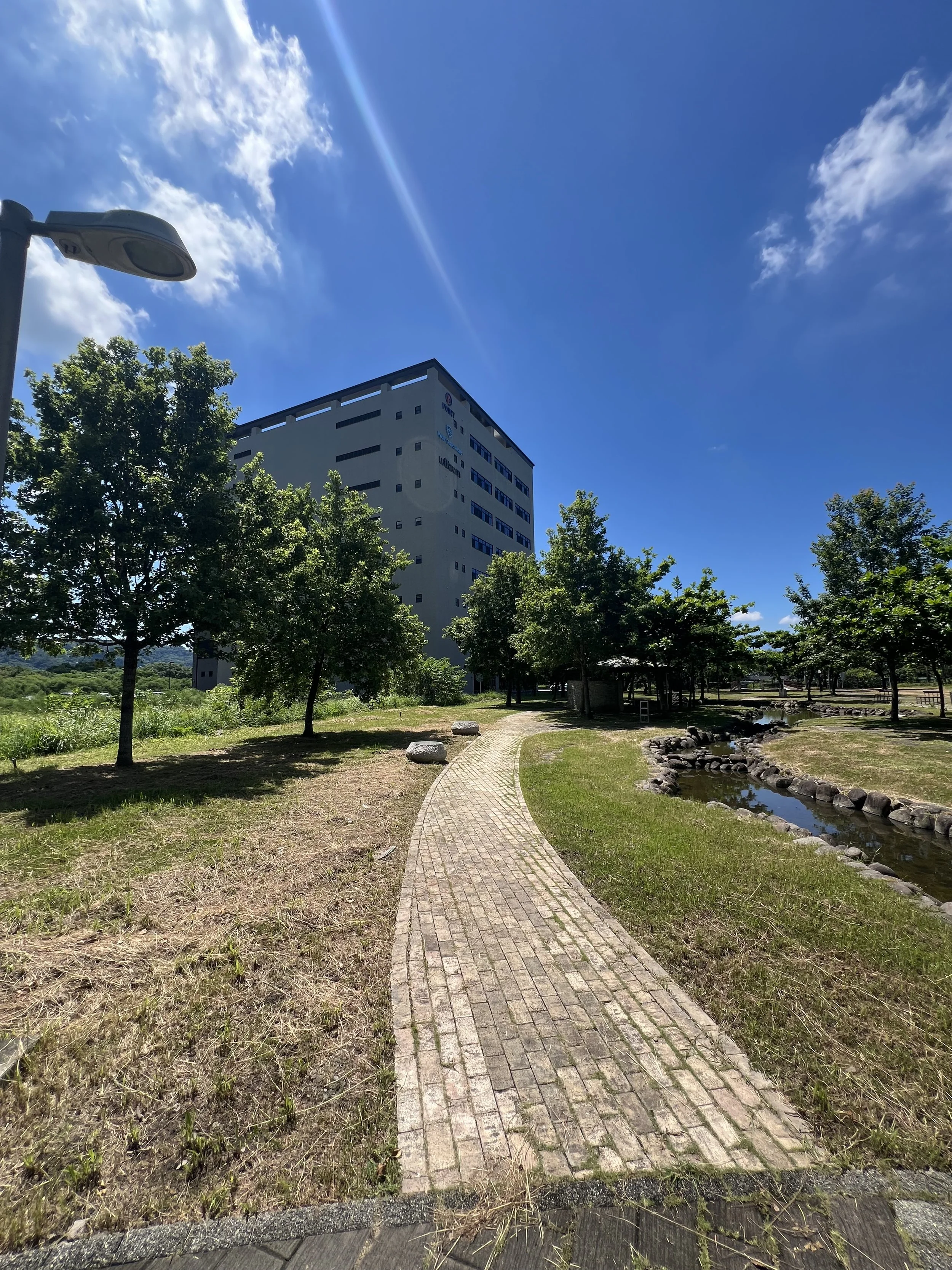 A walking path through a park with trees, a small stream, and a tall building under a partly cloudy blue sky.