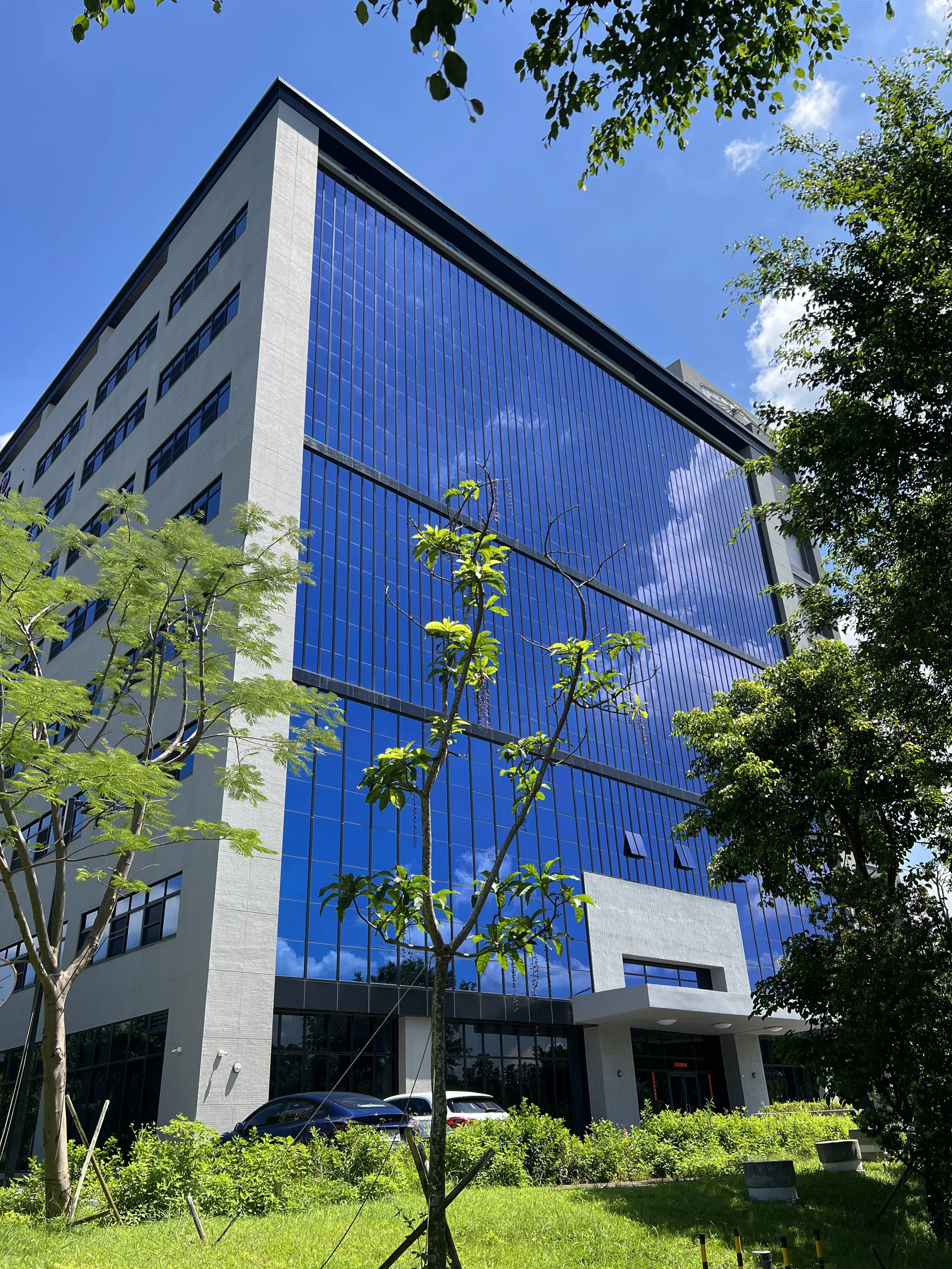 A modern multi-story office building with a glass facade reflecting the blue sky and white clouds, surrounded by green trees and bushes in the foreground.