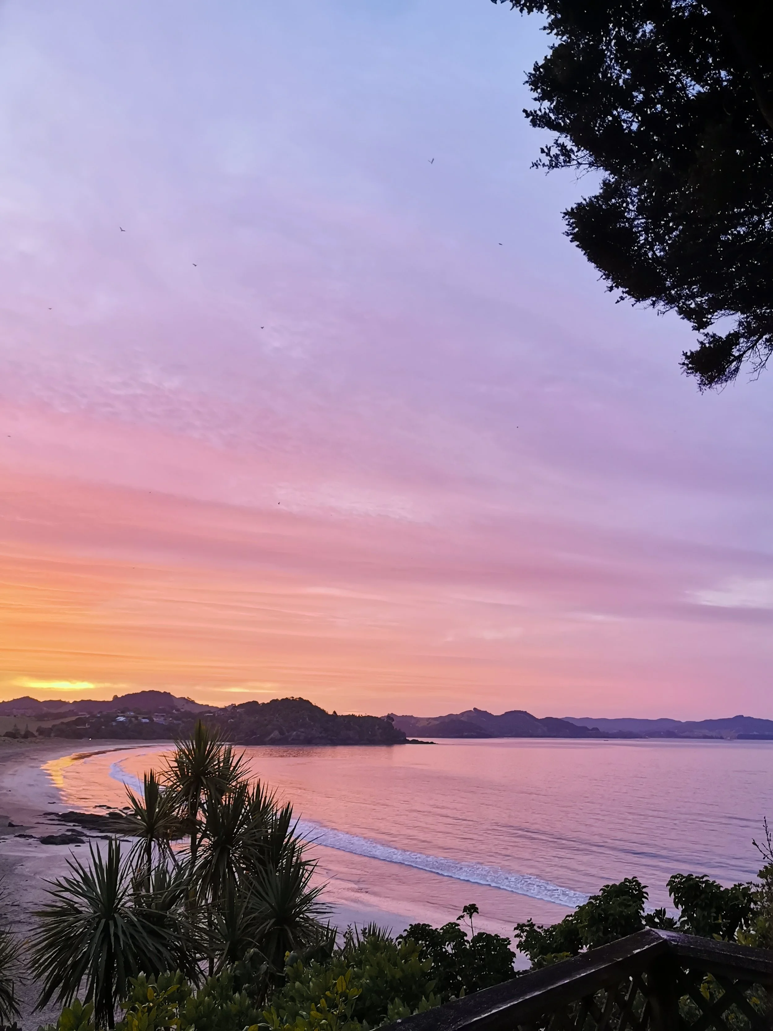 A scenic view of a beach at sunset with colorful sky, calm waters, and silhouettes of hills and trees in the foreground.