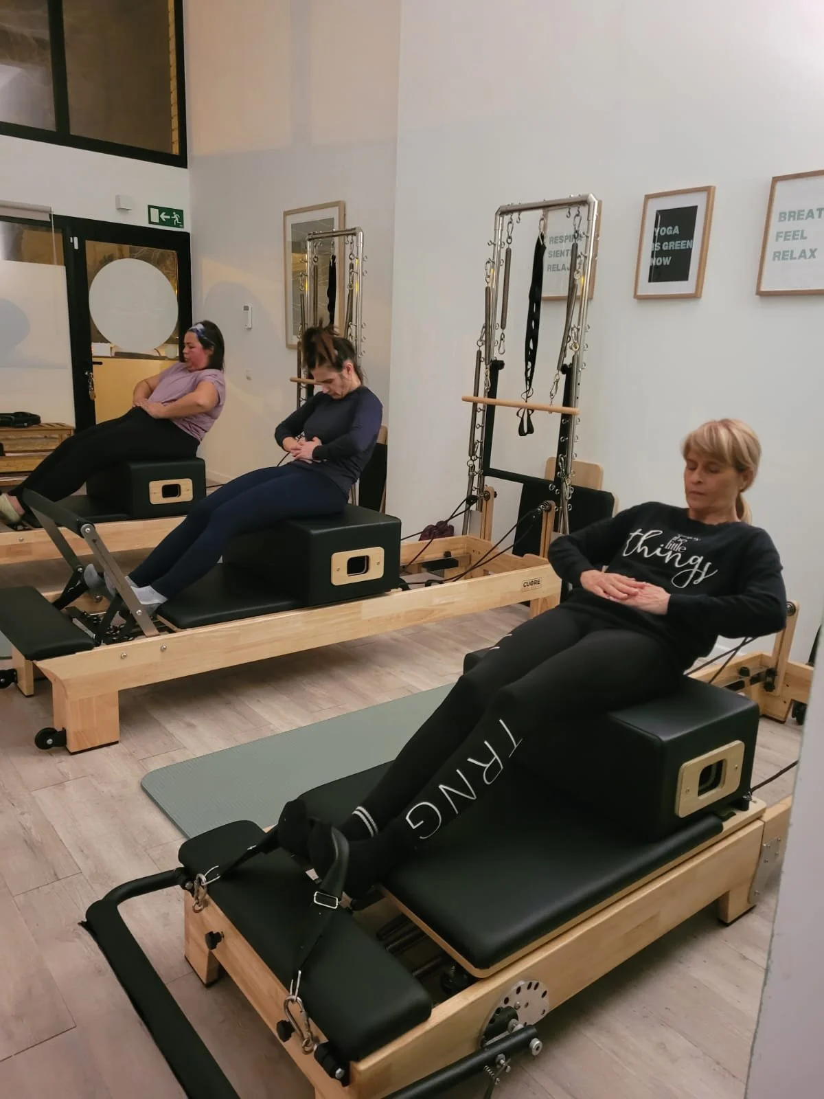 Tres alumnas de Alma Studio Palmanova, cada una en una cama de Pilates, con equipos de ejercicio y fotos motivacionales en la pared que dicen 'Respira, siéntete, relájate', y 'Yoga es tuyo, ahora, relajado'.