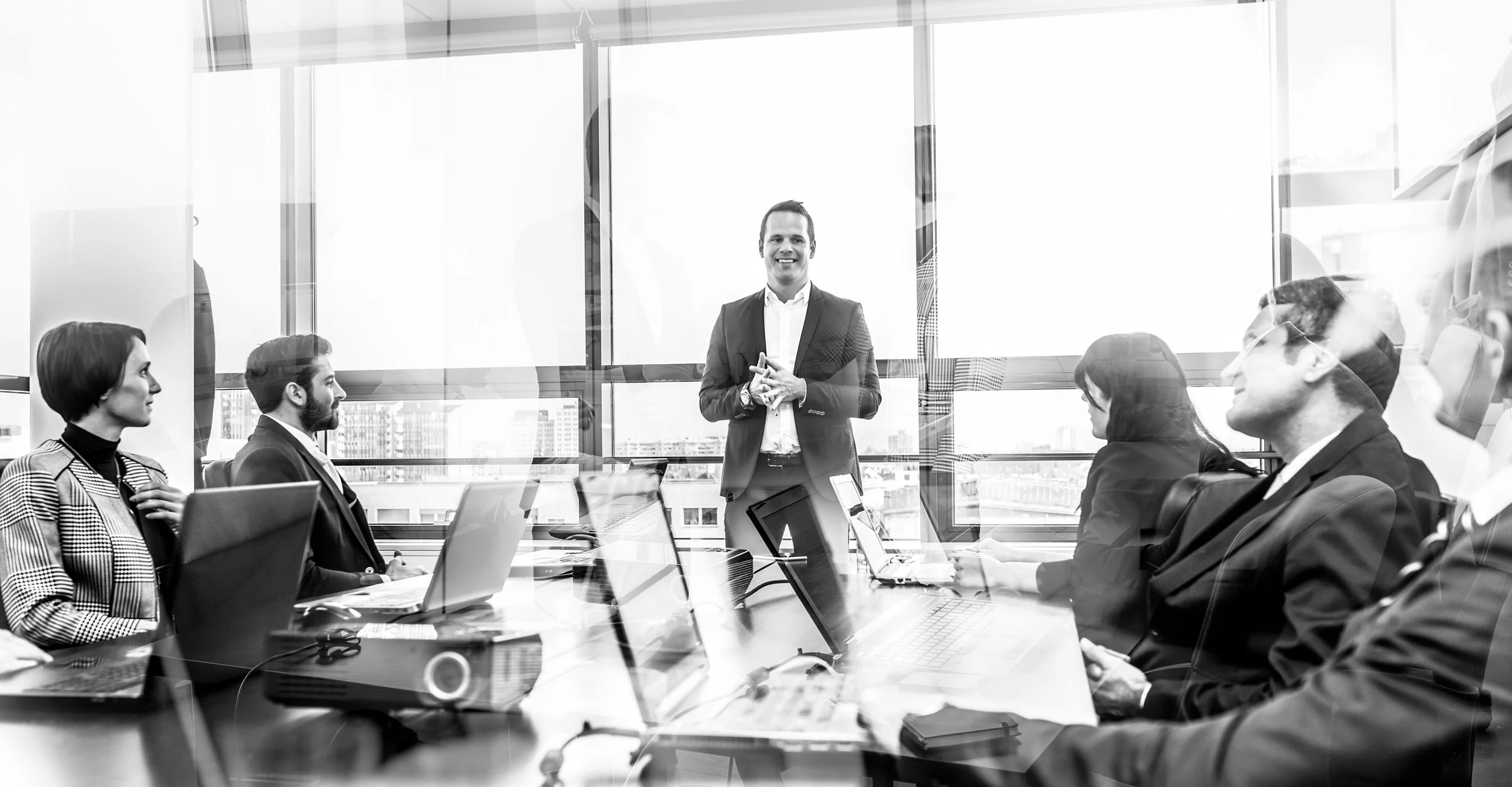 Business meeting in a modern conference room with a presenter standing and smiling, and six seated participants with laptops, facing the presenter.