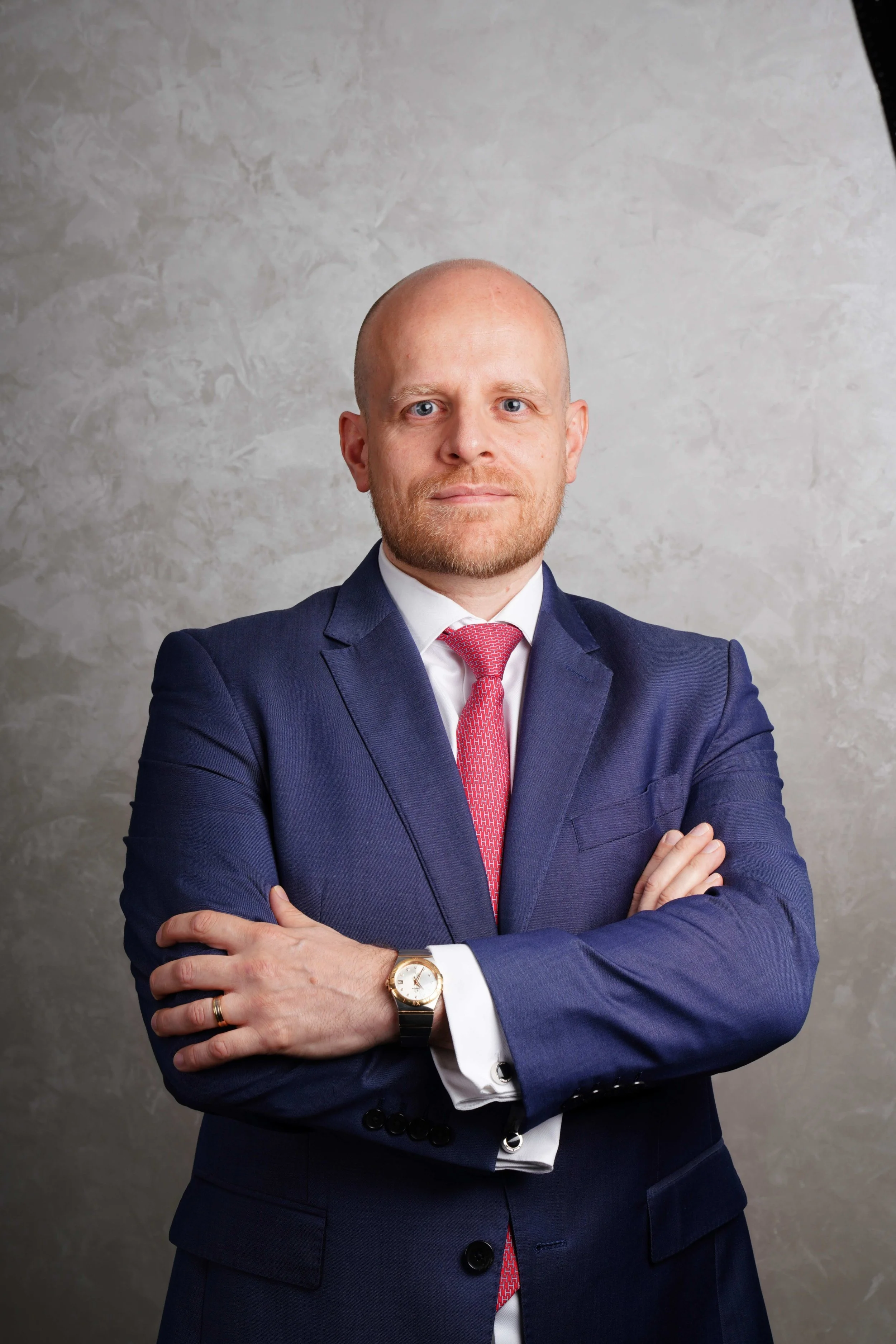 Founder of Kojent Partners, Tim Kroemer, wearing a navy blue suit, white dress shirt, and red tie, crosses his arms and looks at the camera in front of a textured gray background.