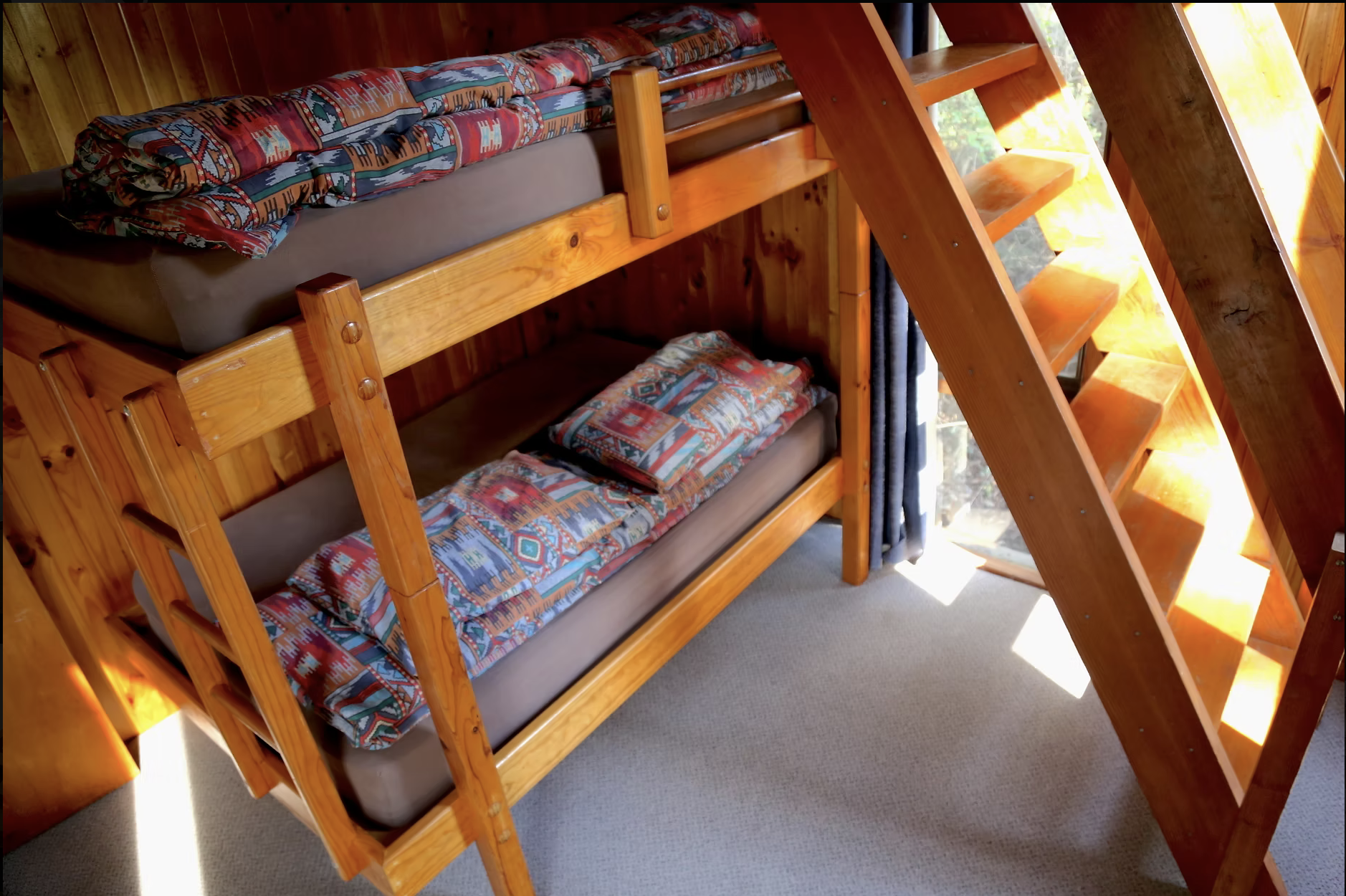 Wooden bunk beds with patterned bedding and pillows, next to a staircase with wooden steps and a window with curtains, in a cozy room with wood paneling walls.