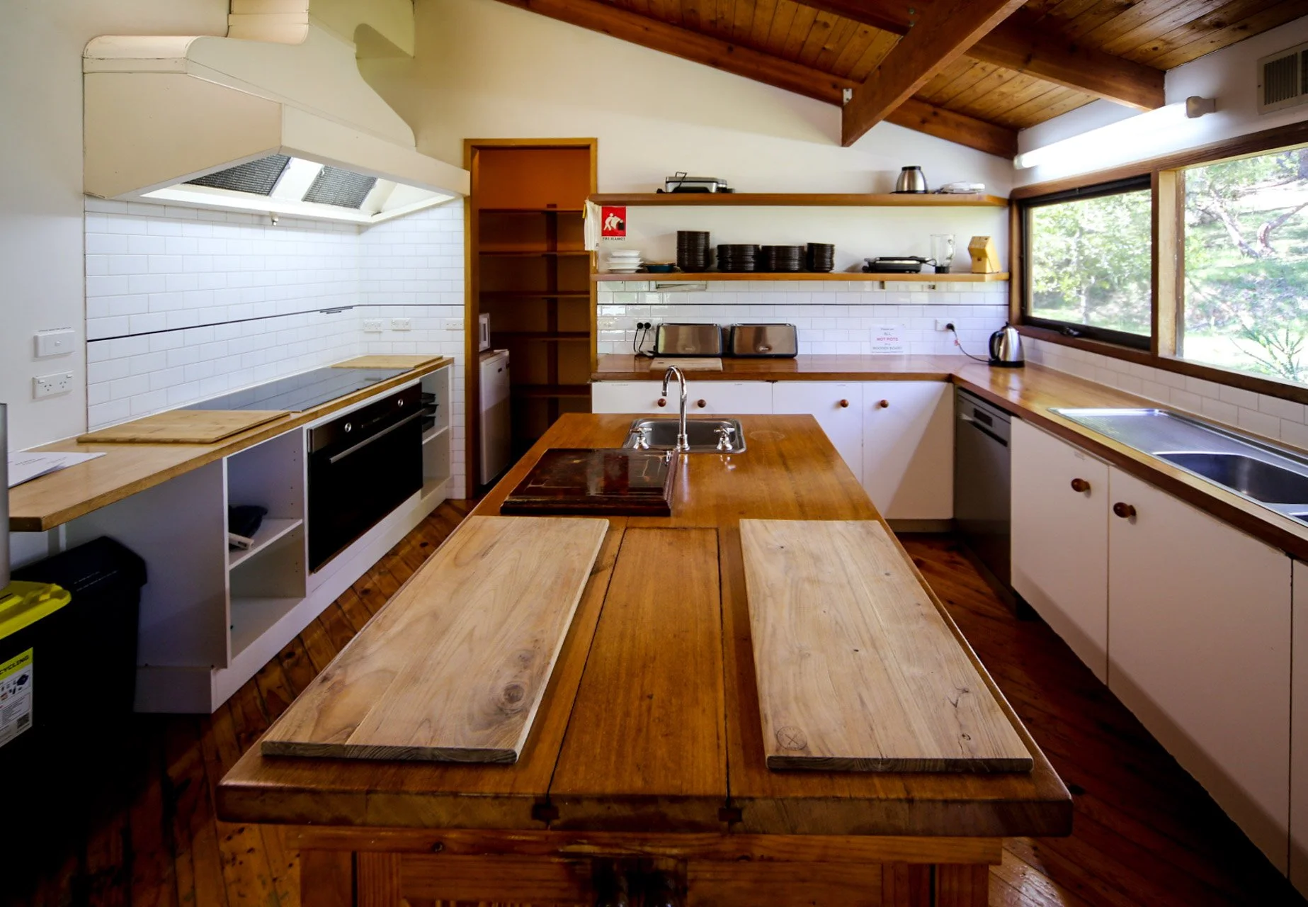 A spacious kitchen with white cabinets, wooden countertops, open shelves with plates and kitchen appliances, a large window overlooking trees, and a wooden dining table in the center.