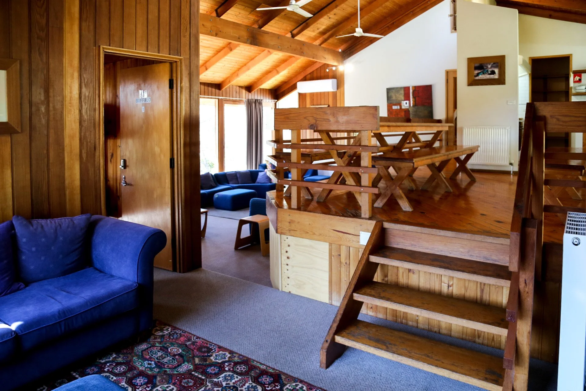 Living room with wooden walls and ceiling, featuring a blue couch, patterned rug, and a raised dining area with wooden tables and benches.