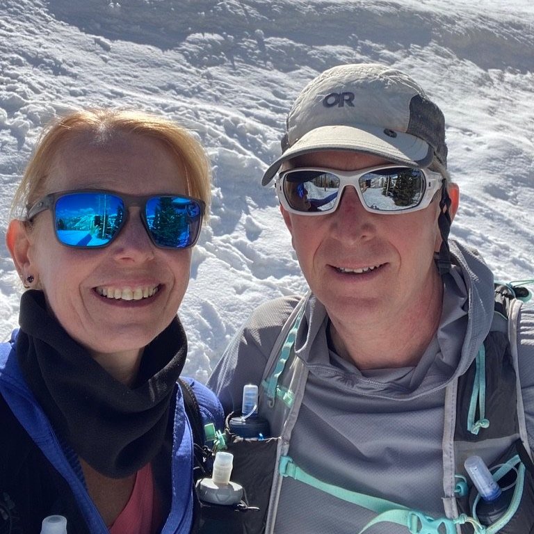 Two smiling hikers in snow, wearing sunglasses and outdoor gear, taking a selfie in a snowy landscape.