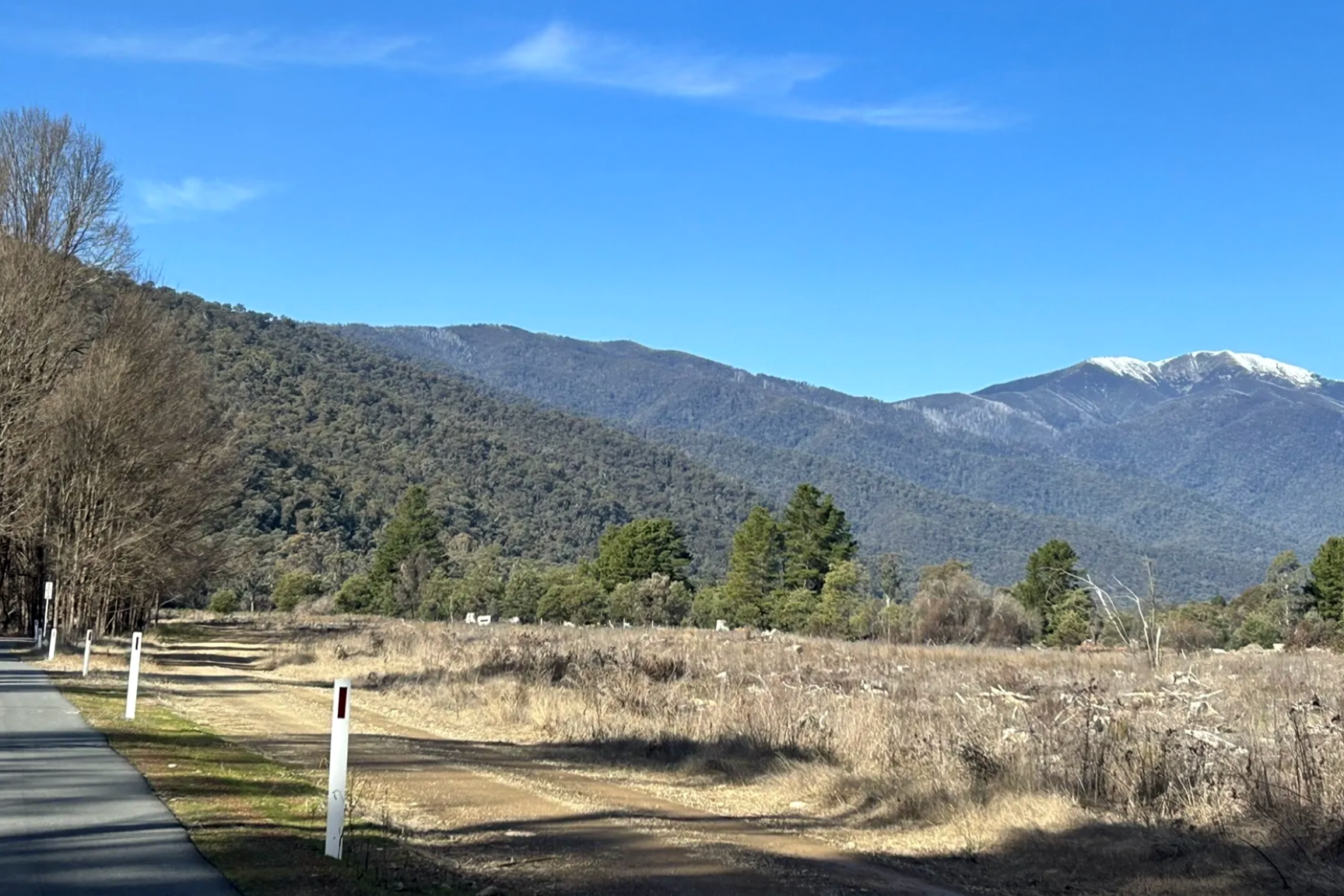 Scenic landscape of the Victorian High Country with snow-capped peaks in the distance, a clear blue sky, trees and bushland, with paved rail trail from Harrietville to Bright in the foreground.