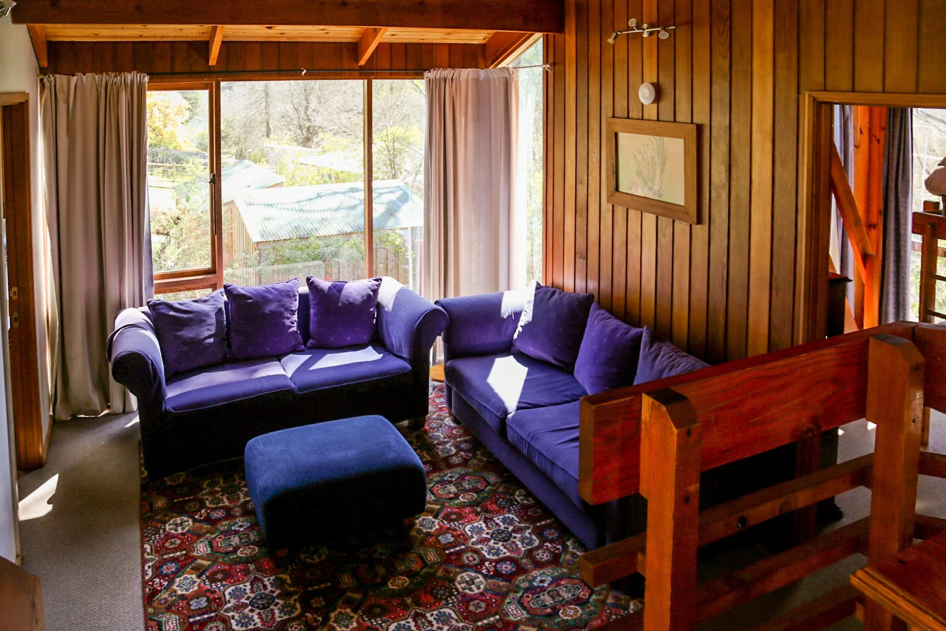 Living room with two purple sofas, a matching ottoman, wooden walls, large windows with white curtains, and a colorful patterned rug.