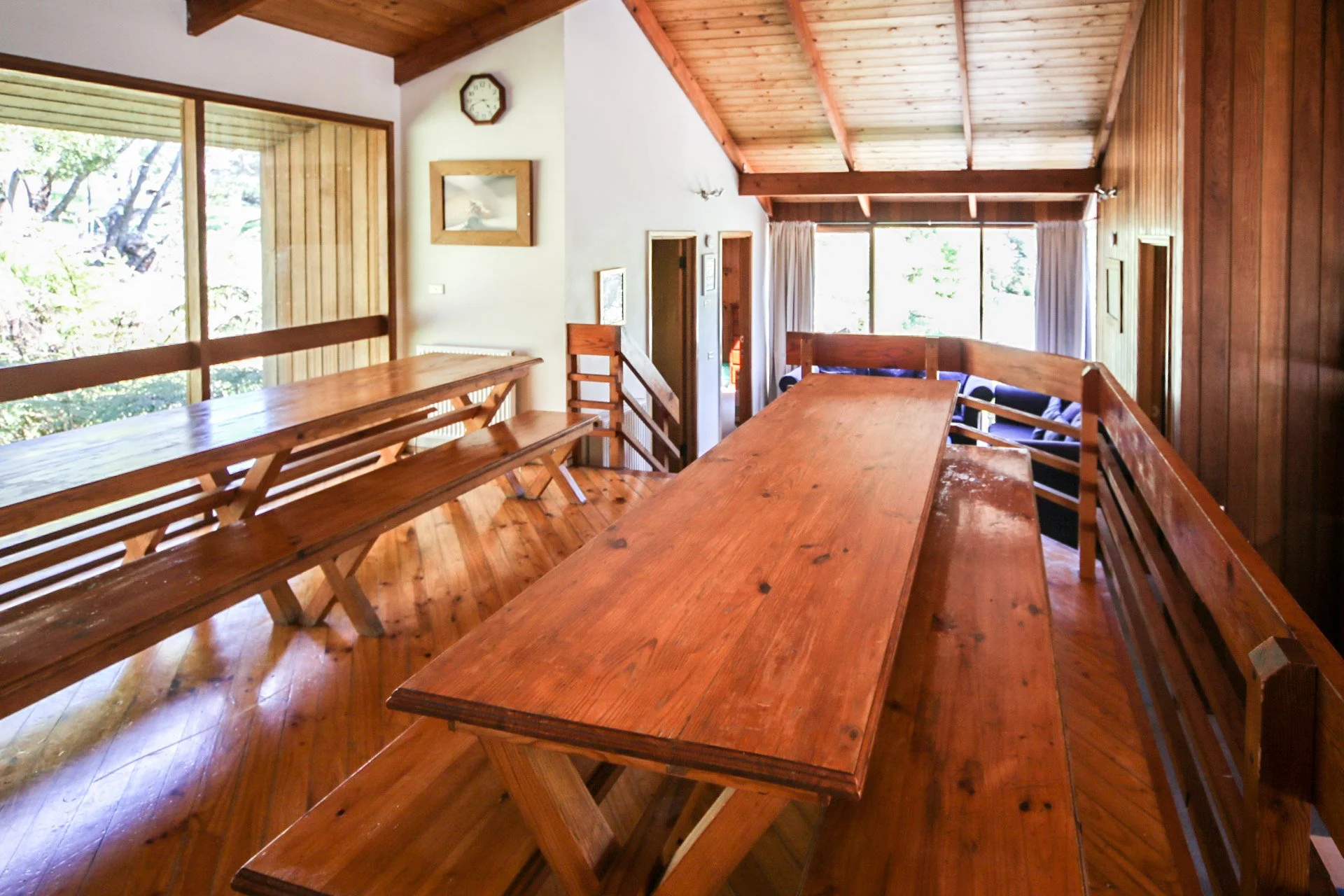 Interior view of a wooden house with large tables and benches, wood-paneled walls, a high ceiling, large windows, and a staircase leading downstairs.