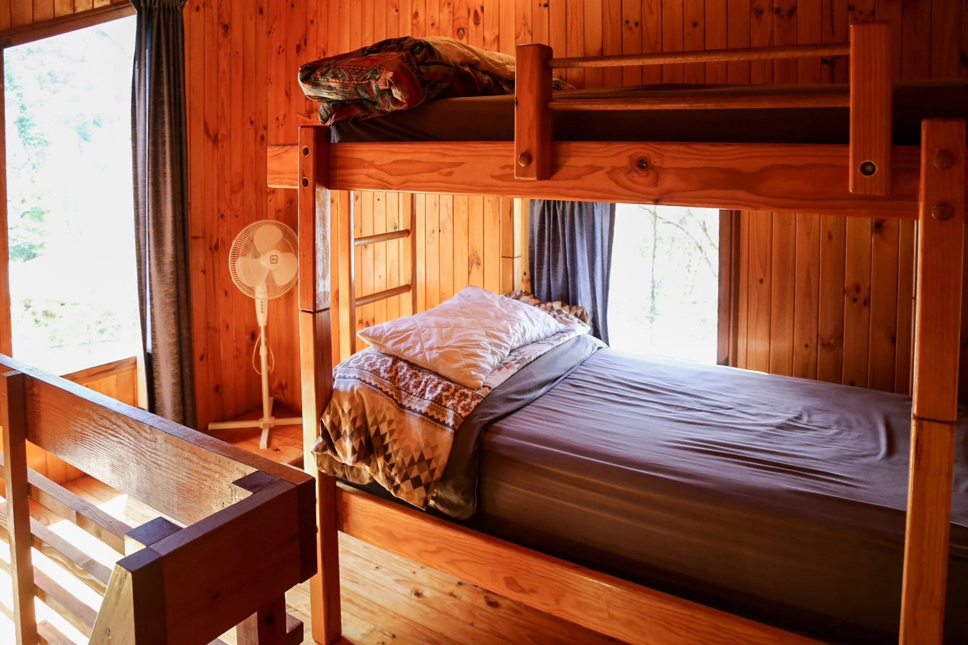 Wood-paneled room with bunk beds, pillows, a small standing fan, and open windows with curtains