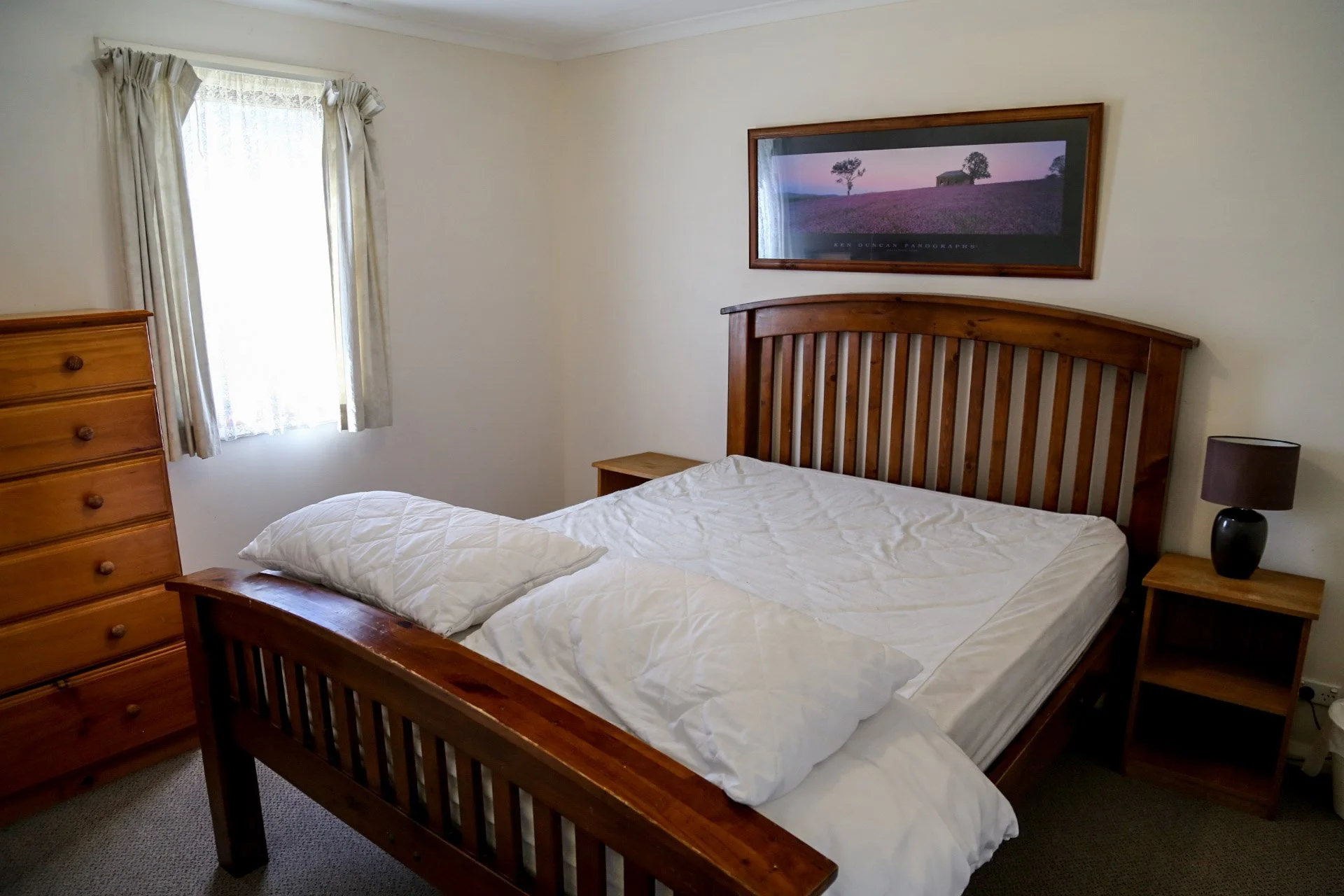 A bedroom with a wooden bed frame and white bedding, a window with cream curtains, a wooden dresser, a small wooden nightstand with a black lamp, and a framed landscape photograph on the wall.