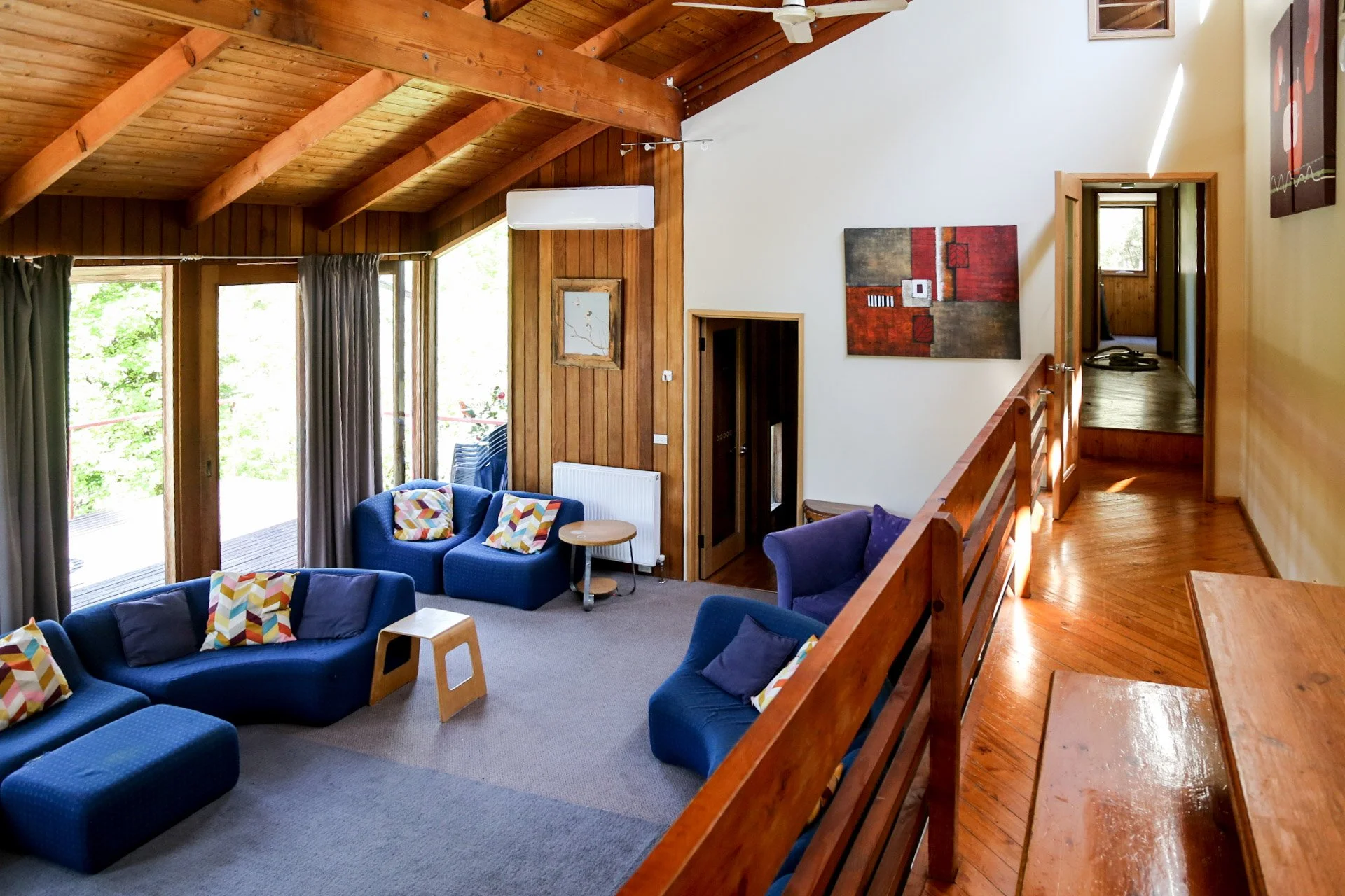 Living room with blue sofas, colorful pillows, wooden ceiling, large sliding doors with curtains, wooden walls, decorative art, and stairs leading to another level.