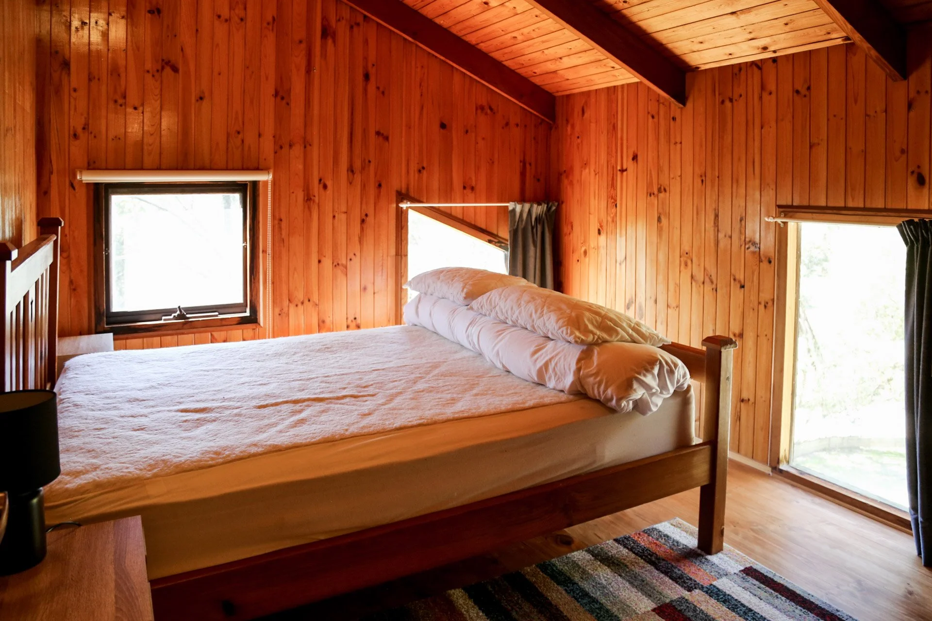 A cozy bedroom with wooden walls and a large bed with white bedding near two windows. One window has a small curtain, and the other has dark curtains. There is a nightstand with a black lamp, and a colorful rug on the wooden floor.