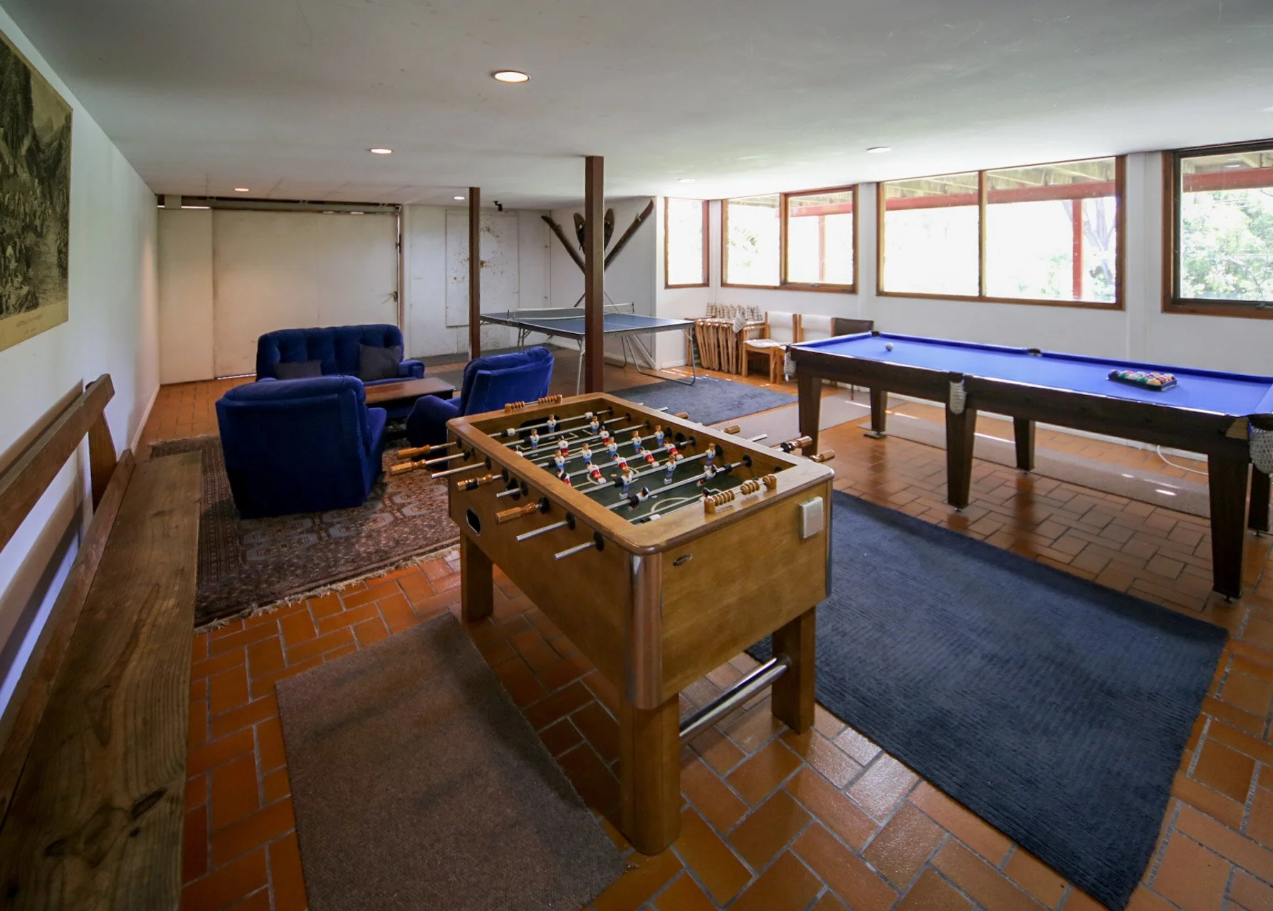 Game room with blue upholstered armchairs, a foosball table, a pool table with a single ball, and a ping pong table, all on brick flooring near large windows.