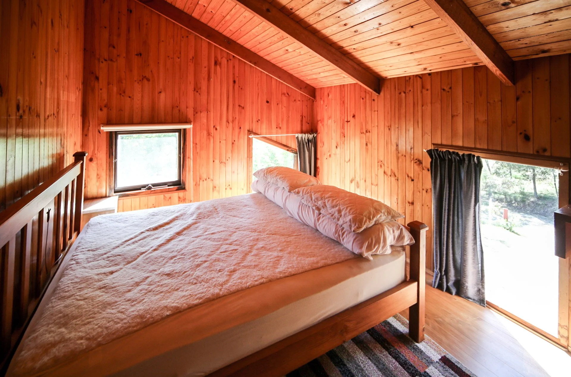 Interior of a cozy bedroom with wood-paneled walls, a large bed with white bedding and pillows, and windows with dark curtains letting in natural light.