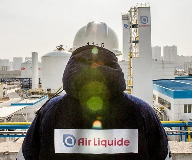 Employé d’Air Liquide portant un casque et une veste marquée du logo, devant une usine industrielle.