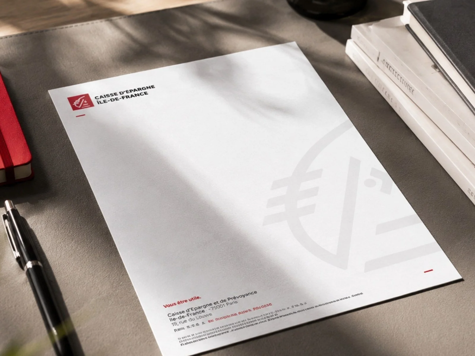 Enveloppe de la Caisse d’Épargne Île-de-France posée sur un bureau, entourée d’un stylo, d’un carnet rouge et de livres.
