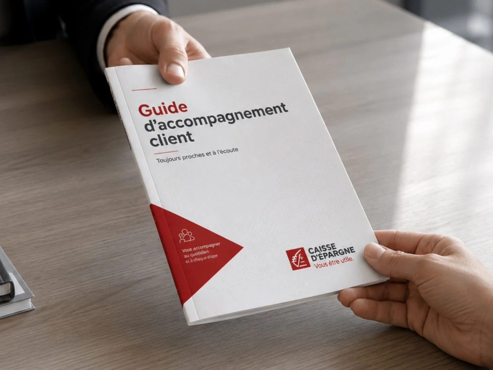 Deux mains tenant un guide d’accompagnement client de la Caisse d’Épargne sur un bureau.