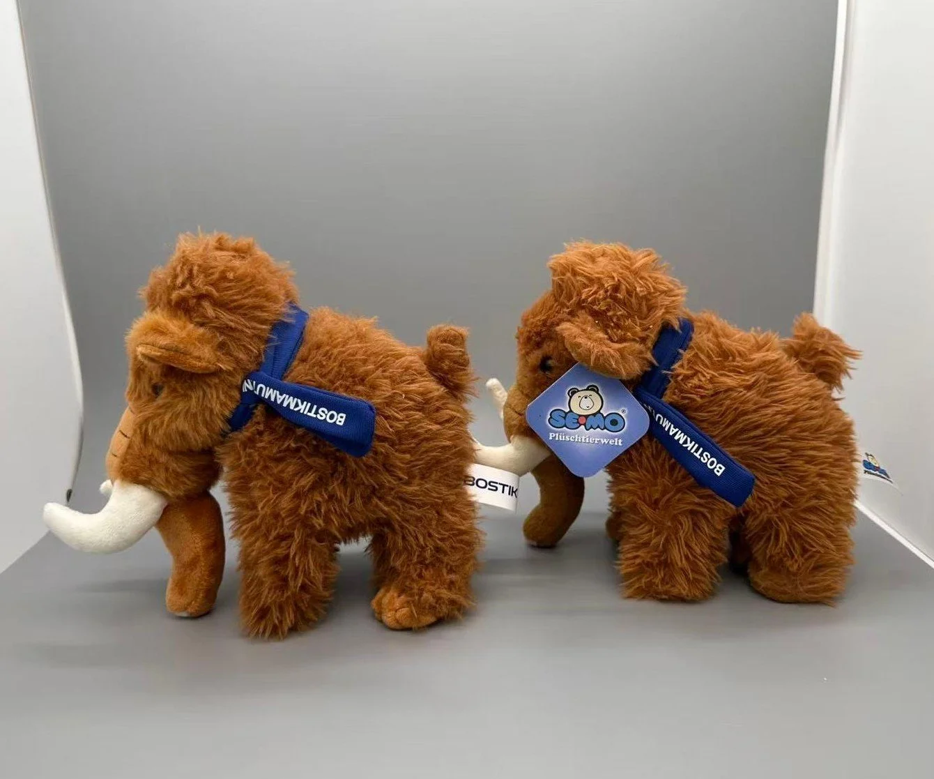 Deux peluches de mammouth brunes avec défenses blanches et étiquettes BOSTIK, présentées sur un fond gris.