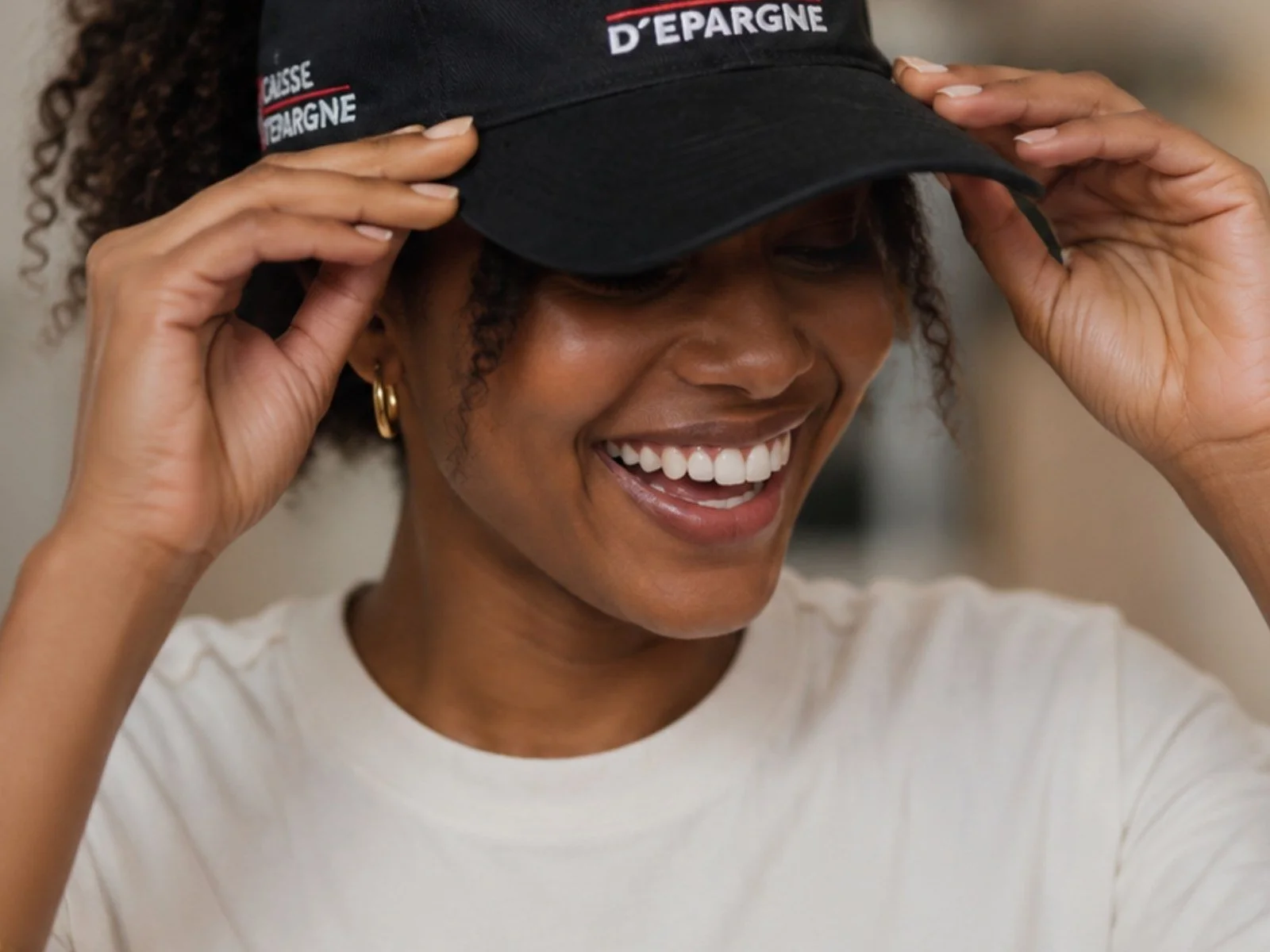 Femme souriante ajustant une casquette noire portant l’inscription « Caisse d’Épargne ».