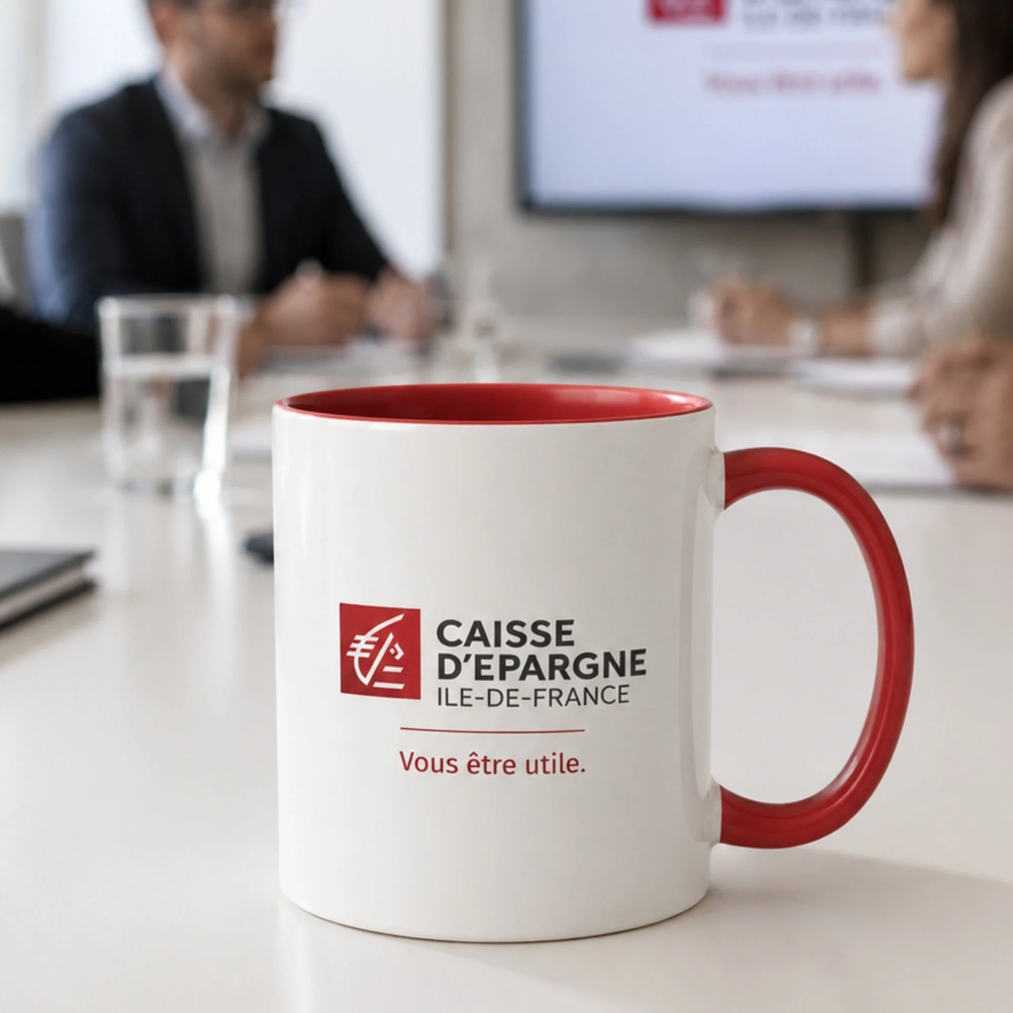 Tasse blanche à bord et anse rouges portant le logo Caisse d’Épargne Île-de-France, posée sur une table de réunion.