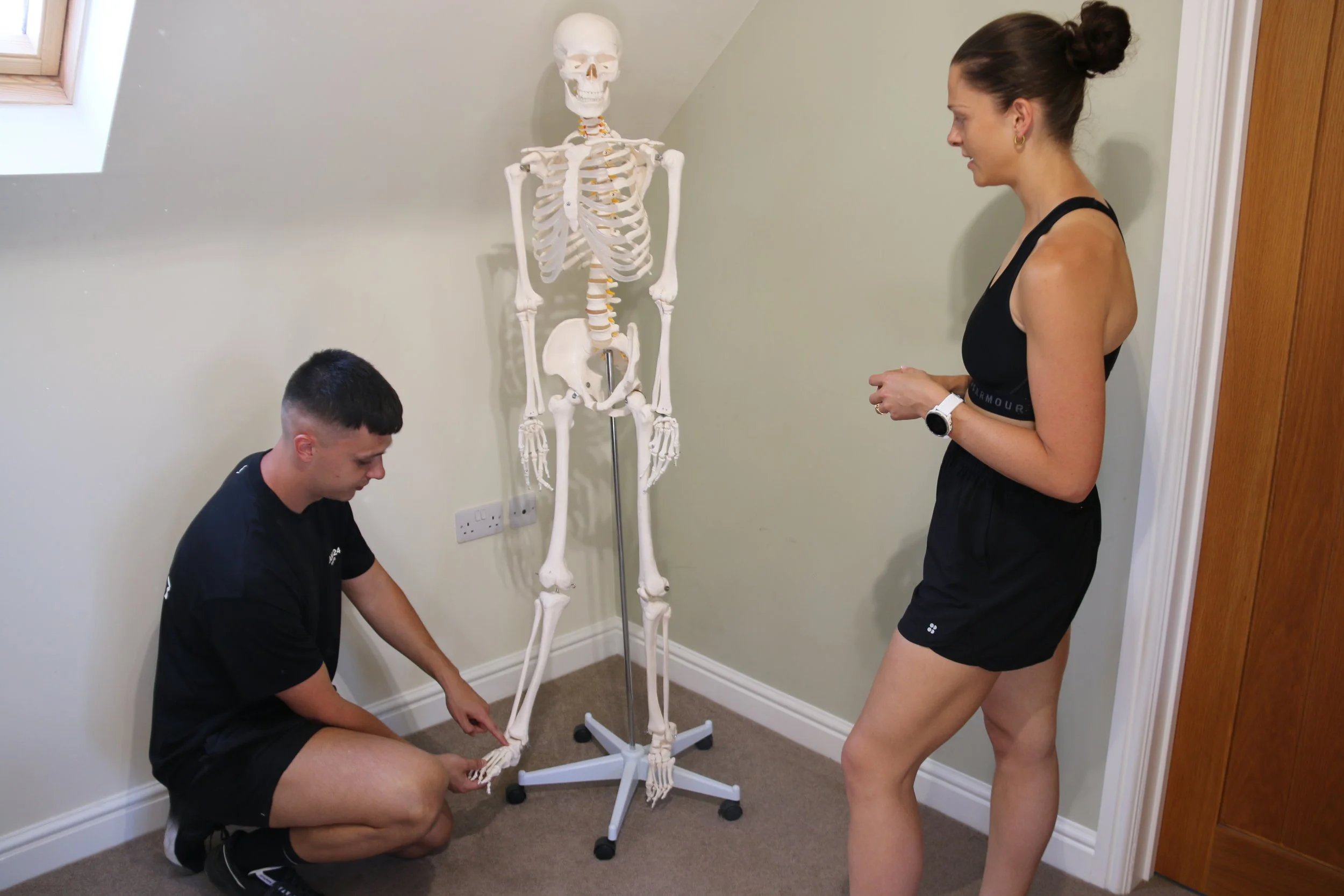A woman and a man examine a human skeleton model in a room with beige walls and a wood door. The woman is standing, wearing a black sports top and shorts, and the man is kneeling, adjusting the skeleton's foot. They are indoors near a window with a skylight.