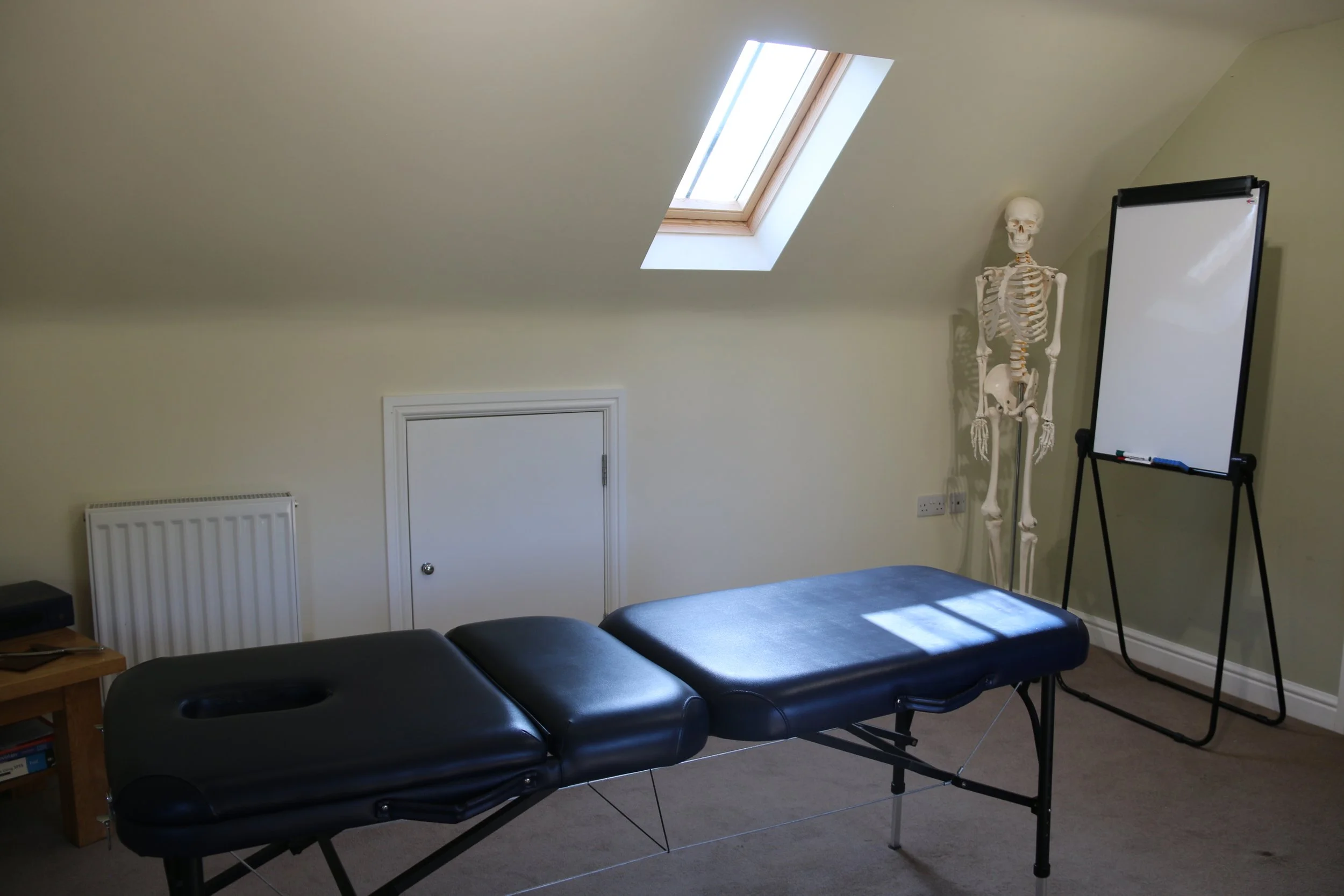 Massage or therapy room with a black massage table, a human skeleton model, a whiteboard on a stand, and a roof window letting in natural light.