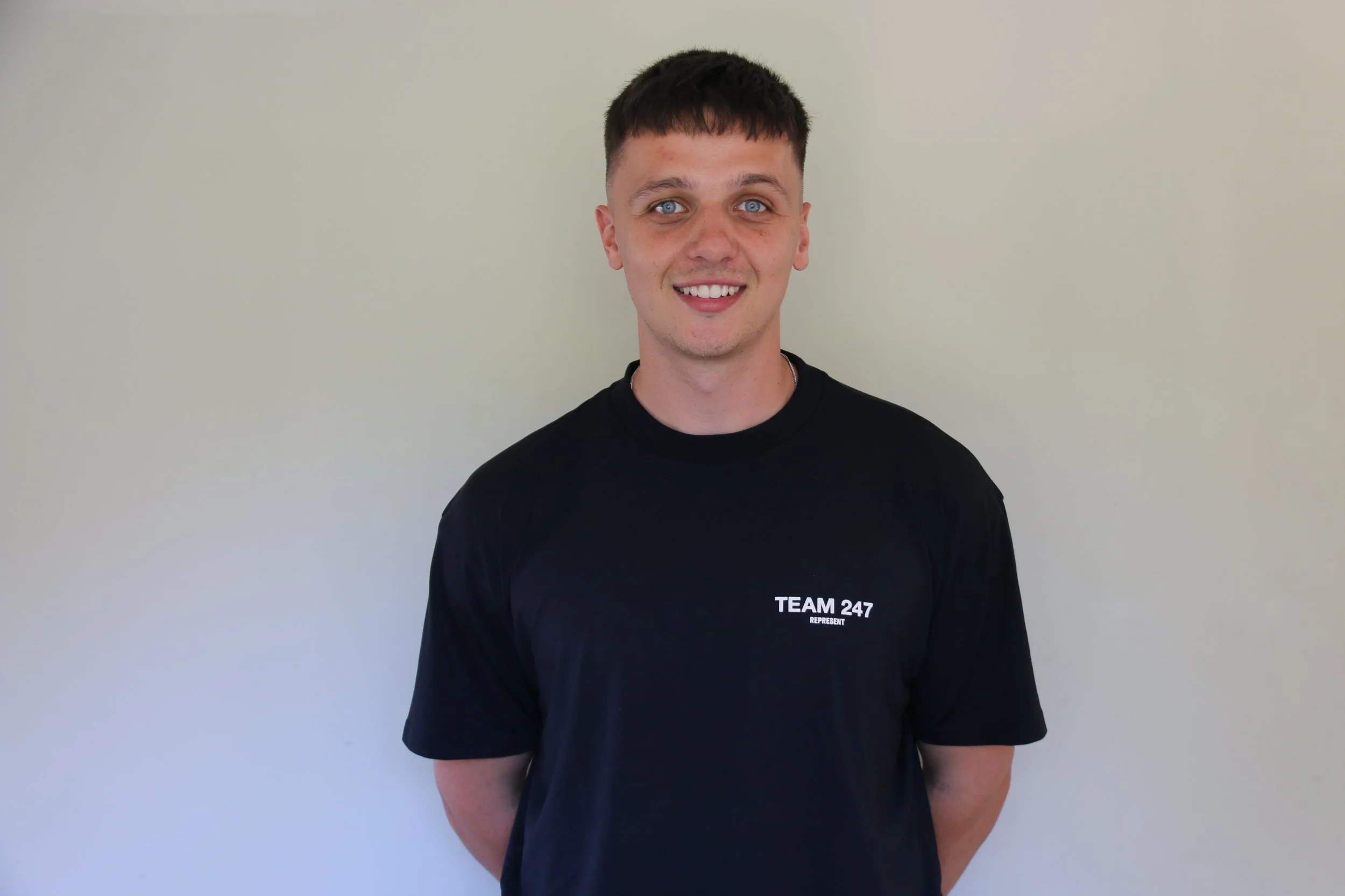 Young man with short brown hair and blue eyes smiling, wearing a black T-shirt with white text that reads 'TEAM 247' standing against a light-colored wall.