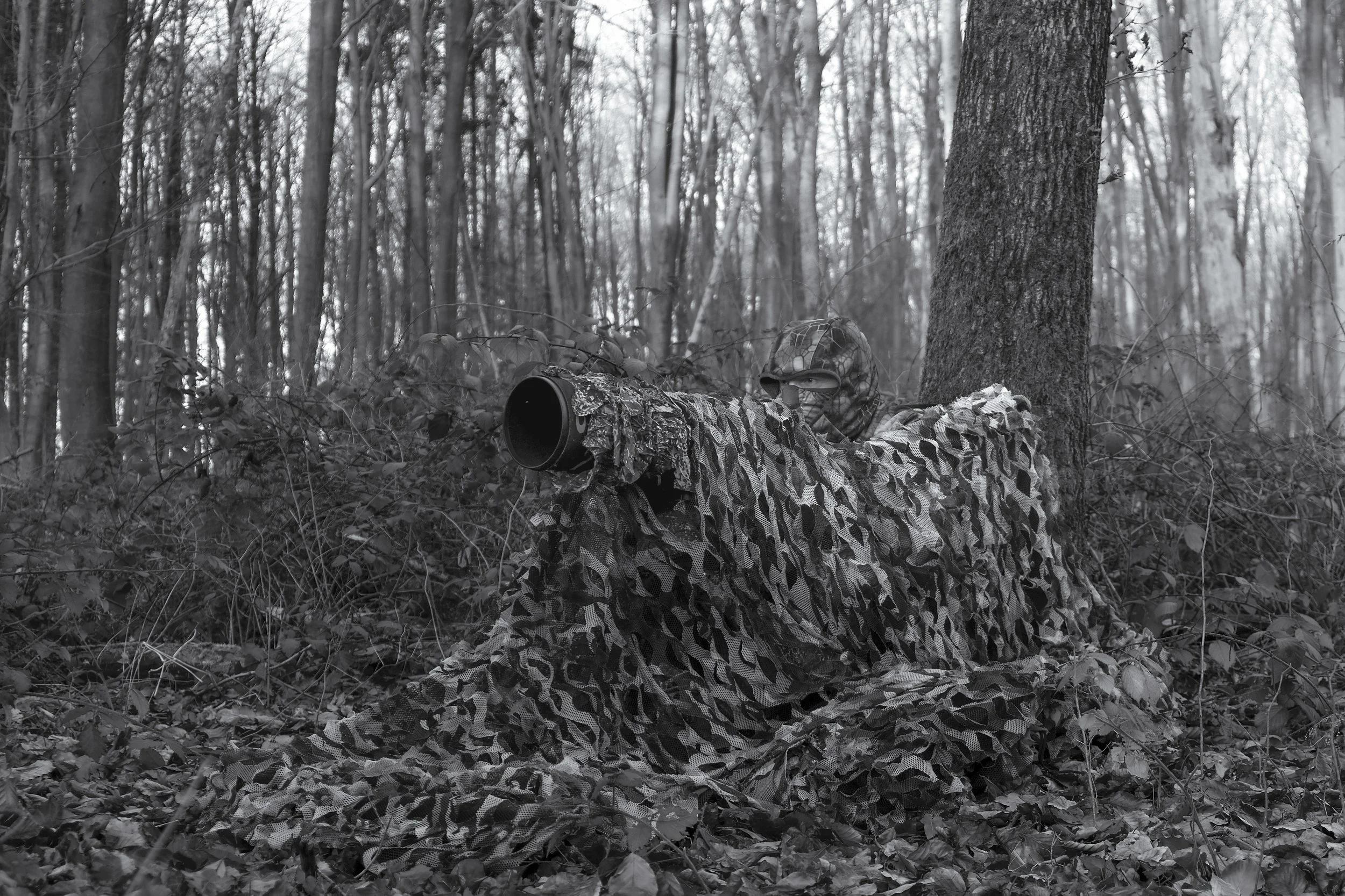 Fotograf in Tarnkleidung, mit einem Tarnnetz, der im Wald eine Beobachtungsposition einnimmt, während die Sonne untergeht. Bild in Schwarz-Weiß.